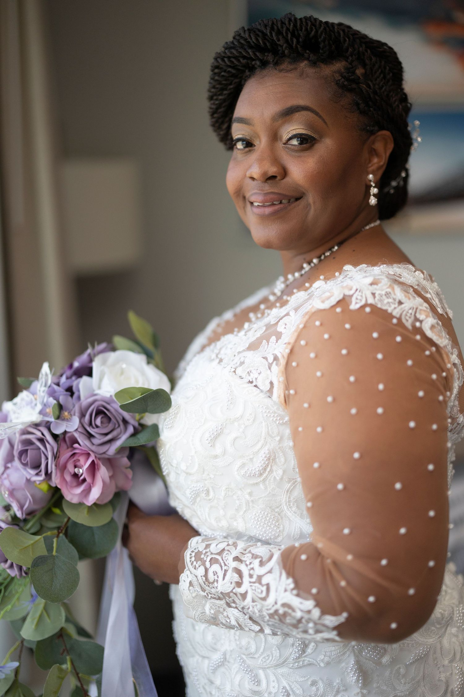 Smiling bride in white lace pearl-embellished gown holding purple and white rose bouquet, portrait photo.