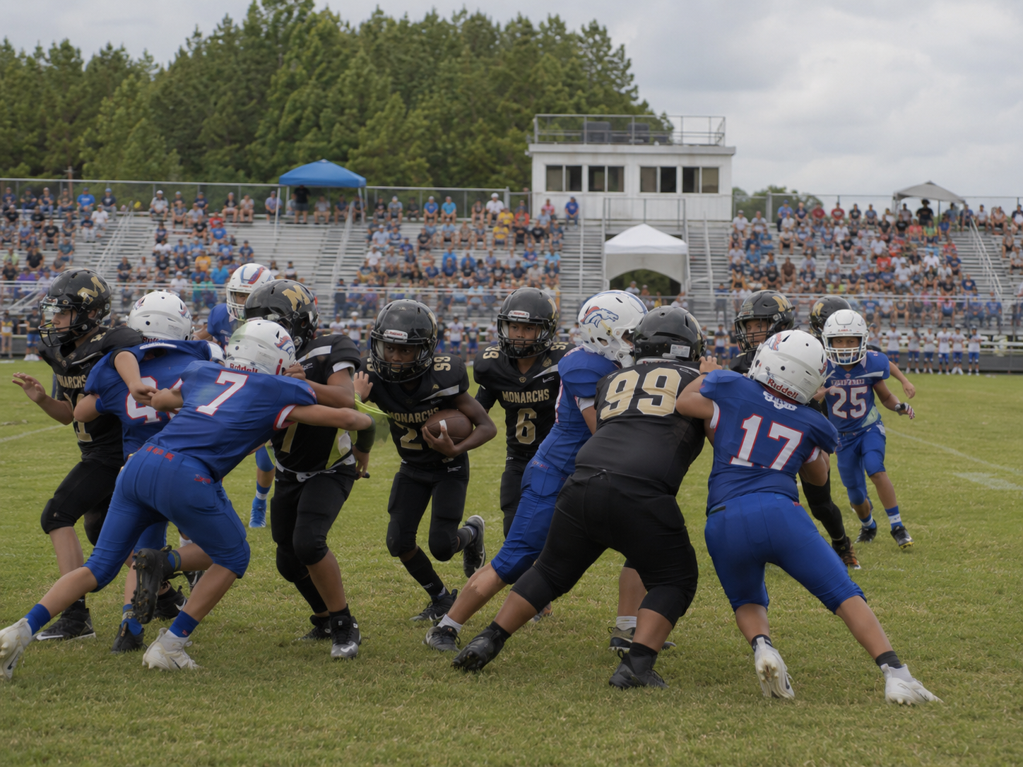 Youth football players clash on a green field during a game with a crowded stadium in the background.