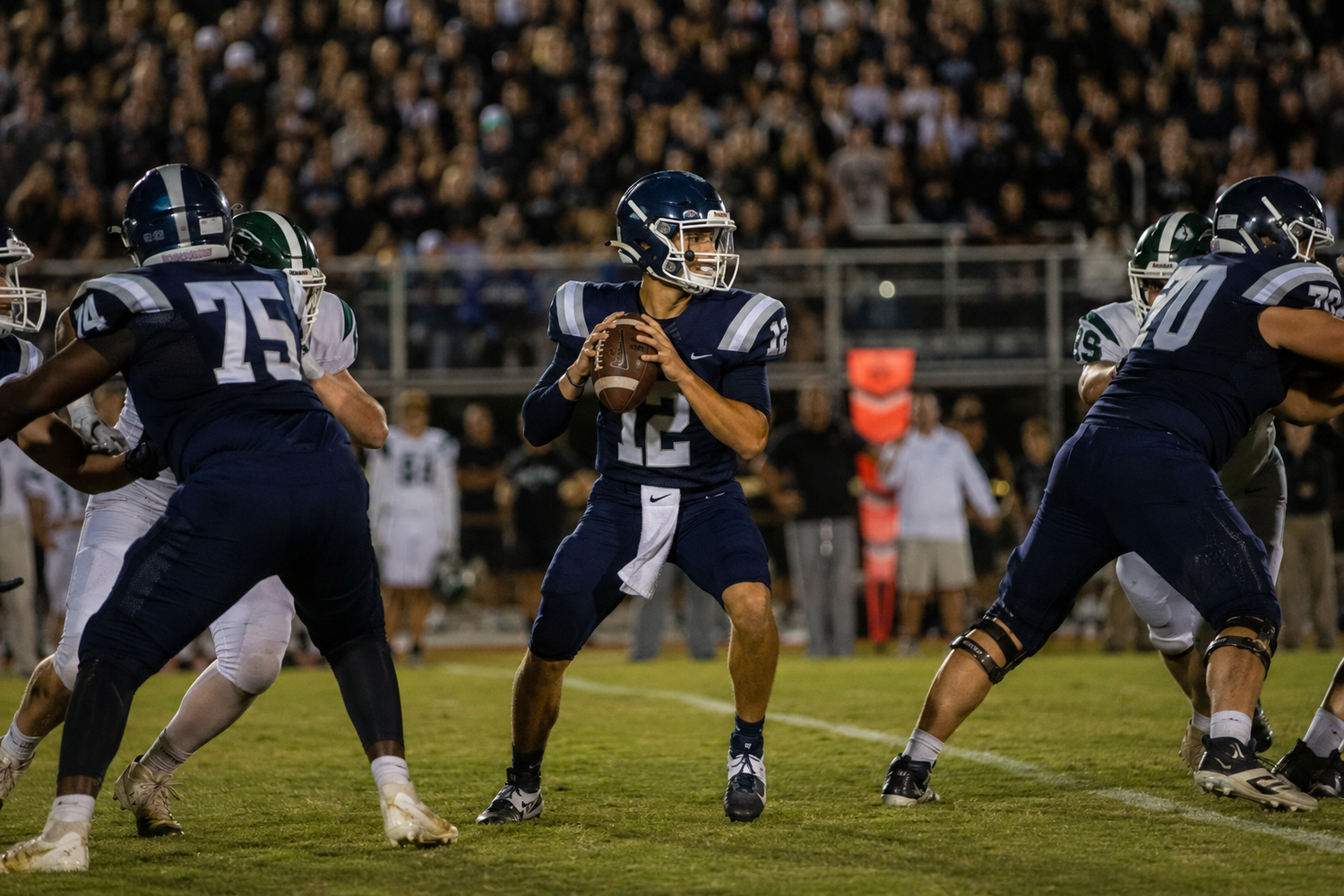 High school football quarterback number 12 drops back to pass under Friday night lights during a competitive game.