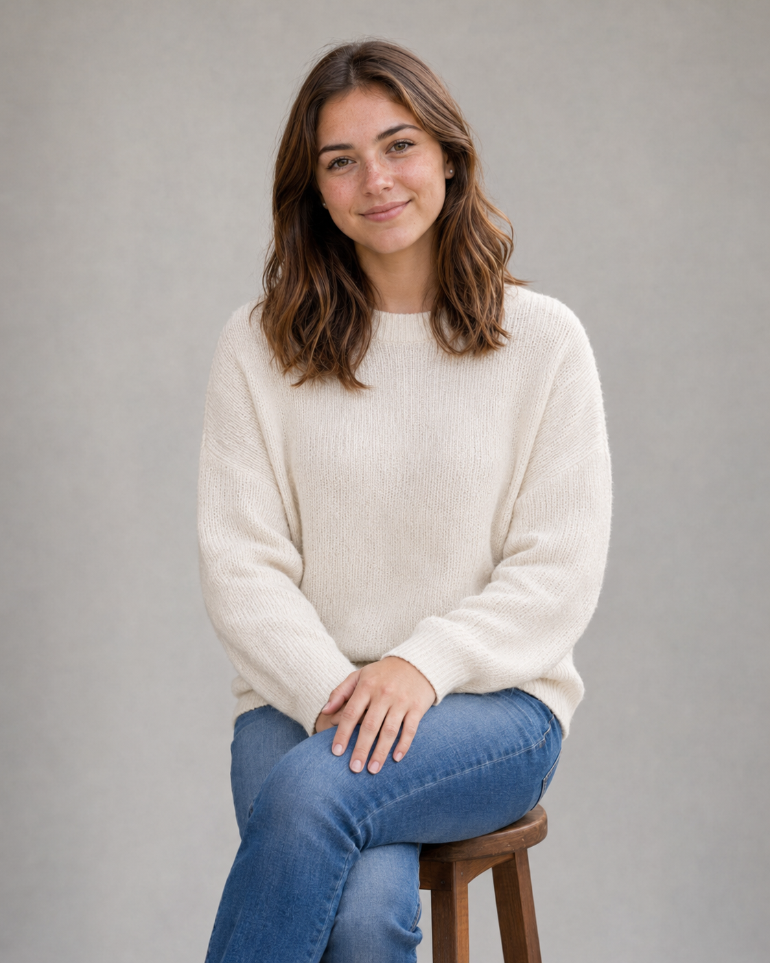 Young woman in cream knit sweater and jeans sitting on a wooden stool against a gray background.