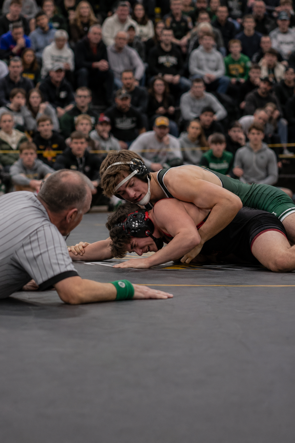 Wrestler in green headgear pins opponent to the mat as referee observes during a crowded wrestling meet.