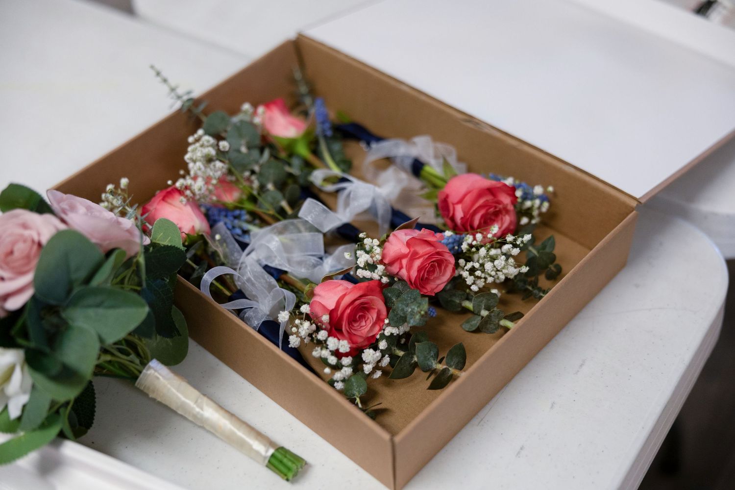A cardboard box containing pink rose boutonnieres with baby's breath and blue ribbon, alongside a floral bouquet on a white surface.