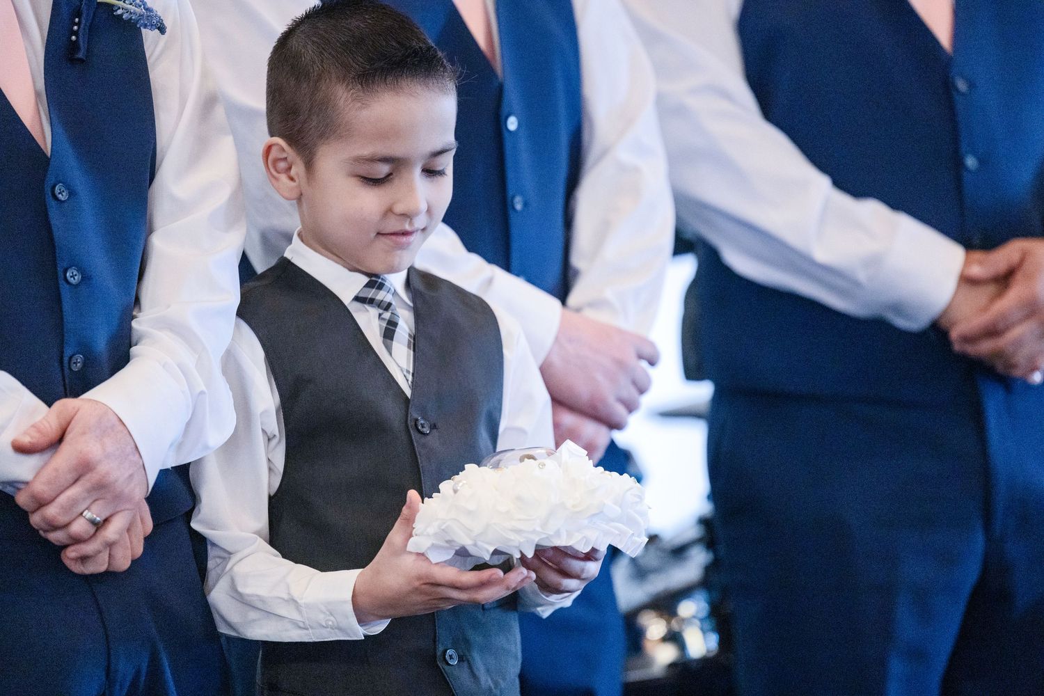 Young ring bearer in grey vest and tie holds wedding rings on white pillow, flanked by groomsmen in navy suits.