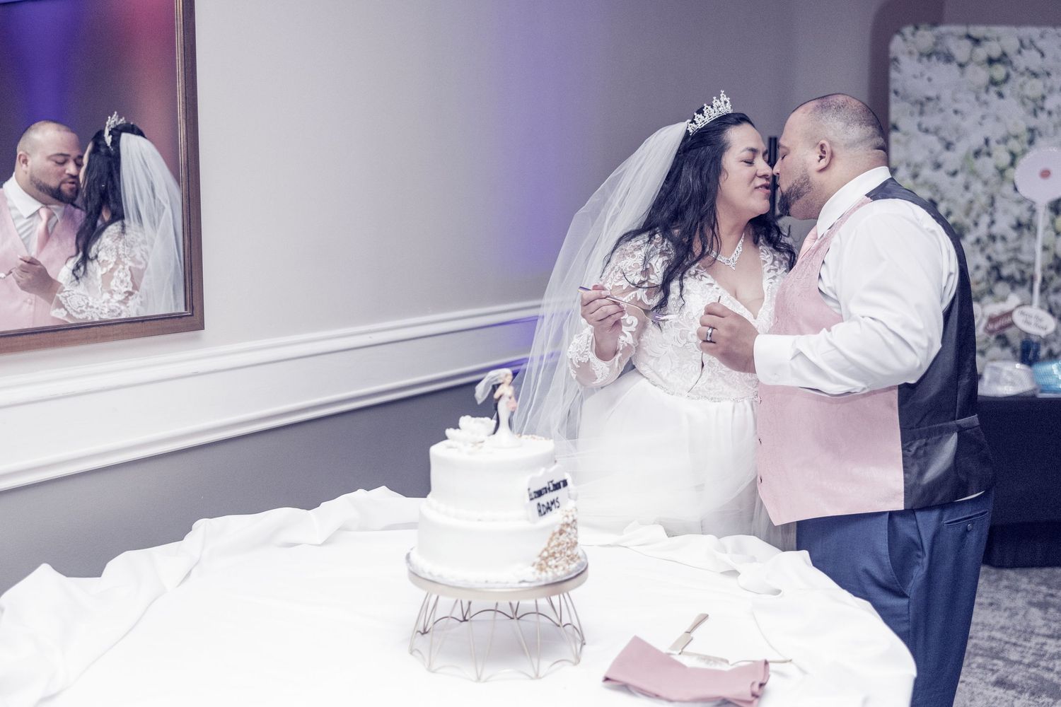 Bride and groom cutting their white wedding cake together, with their reflection visible in a mirror behind them.