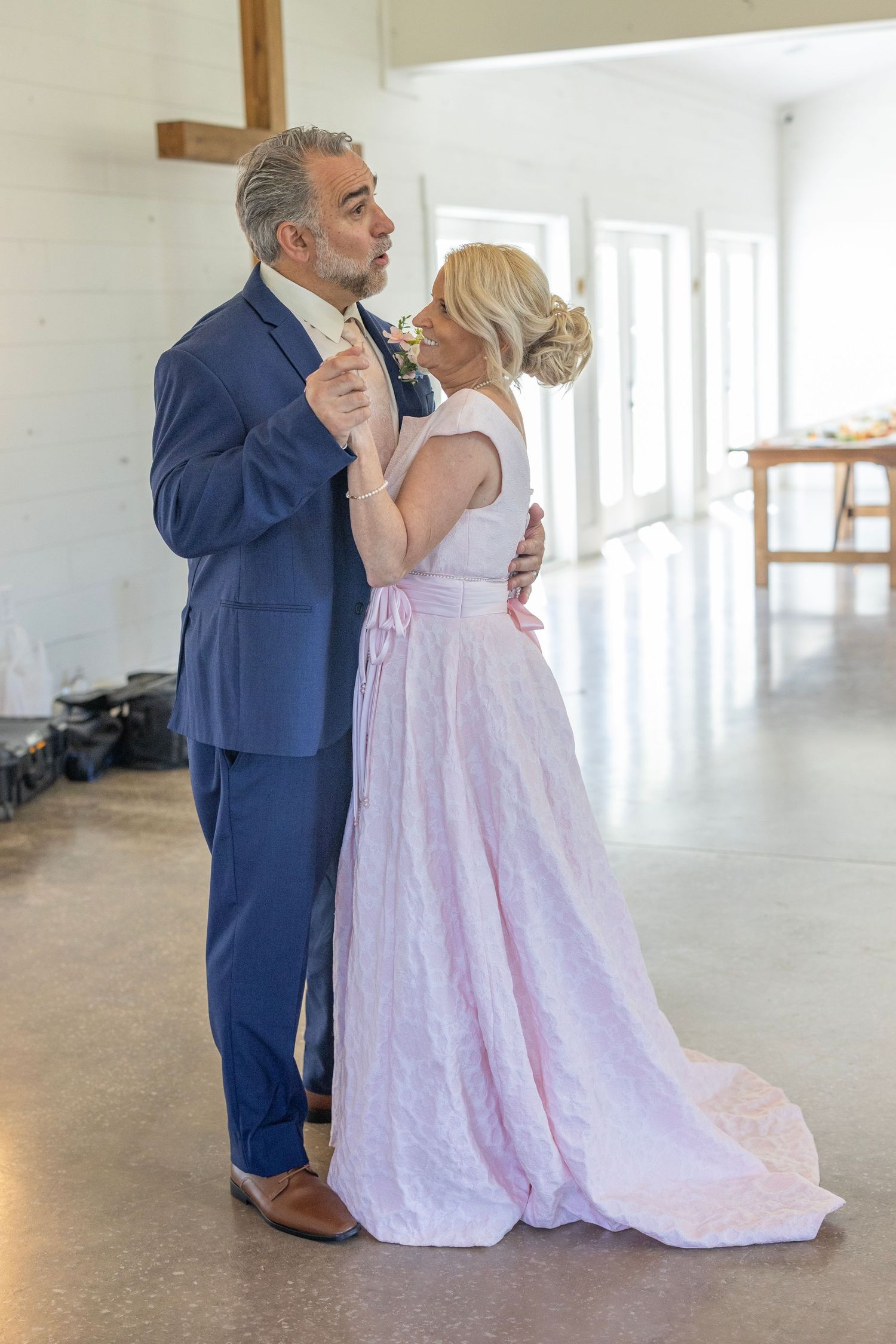 A couple shares their first dance, man in blue suit, woman in pink gown, inside a bright venue with a cross on the wall.