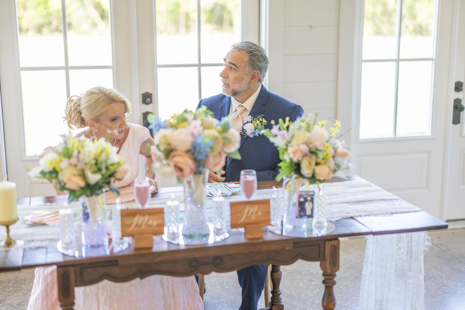 Elegant wedding couple seated at a decorated sweetheart table with pastel floral arrangements in a bright venue.