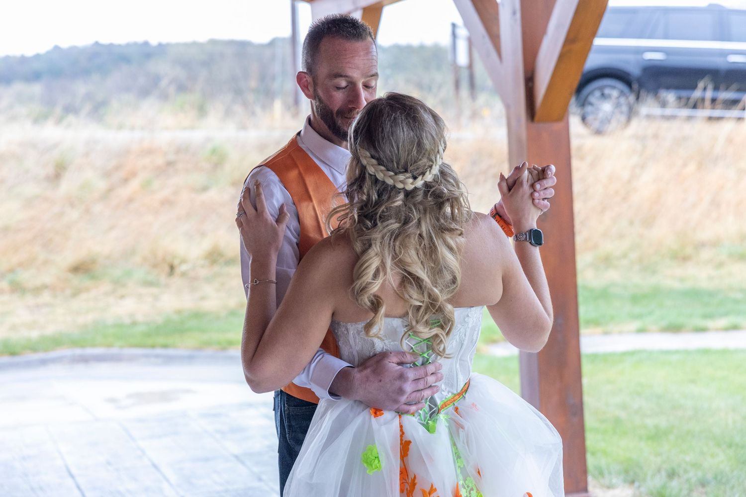 Bride and groom share their first dance outdoors under a wooden pergola, wearing colorful paint-splattered wedding attire.