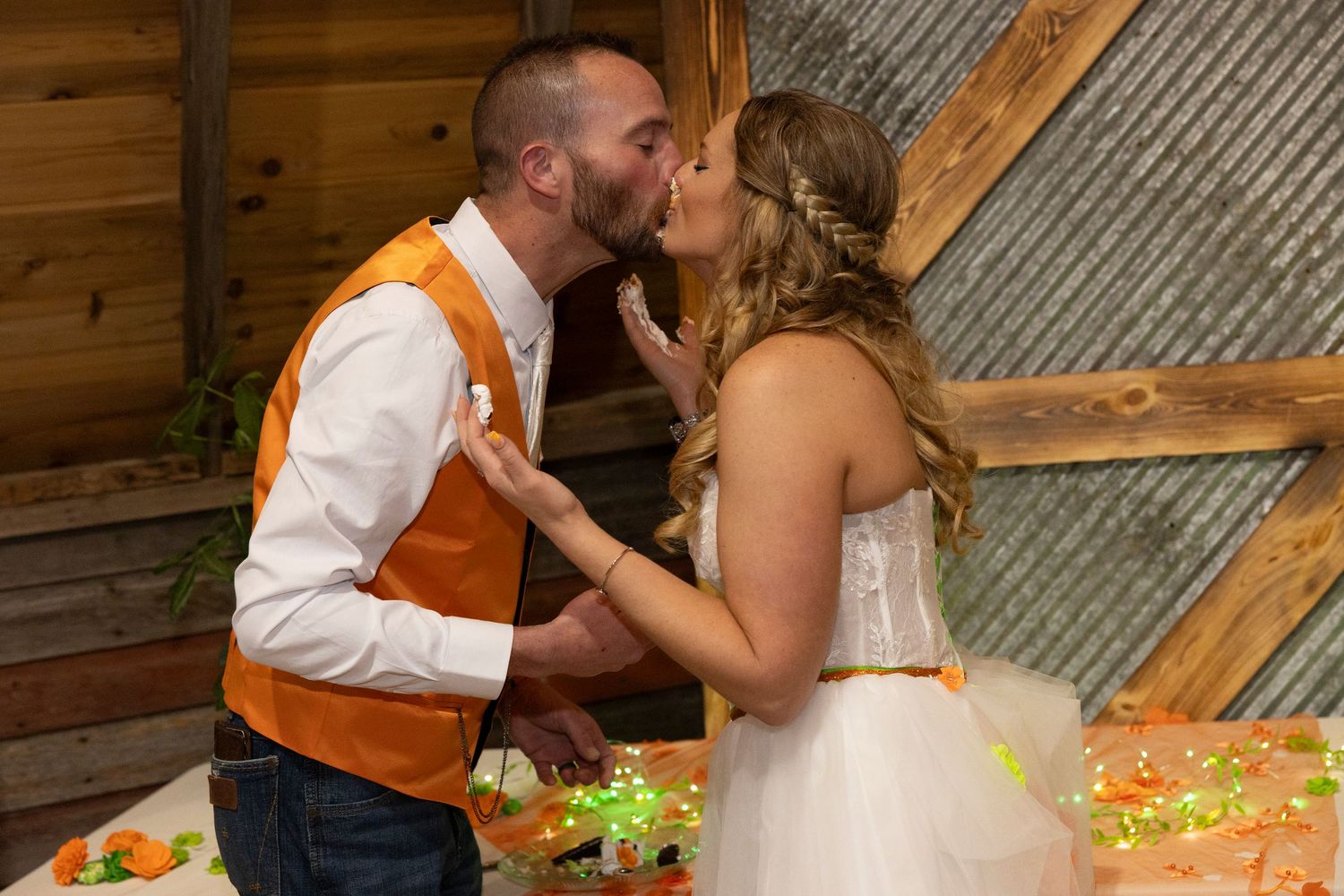 Bride and groom sharing a kiss at rustic barn wedding reception with orange accents and cake table.