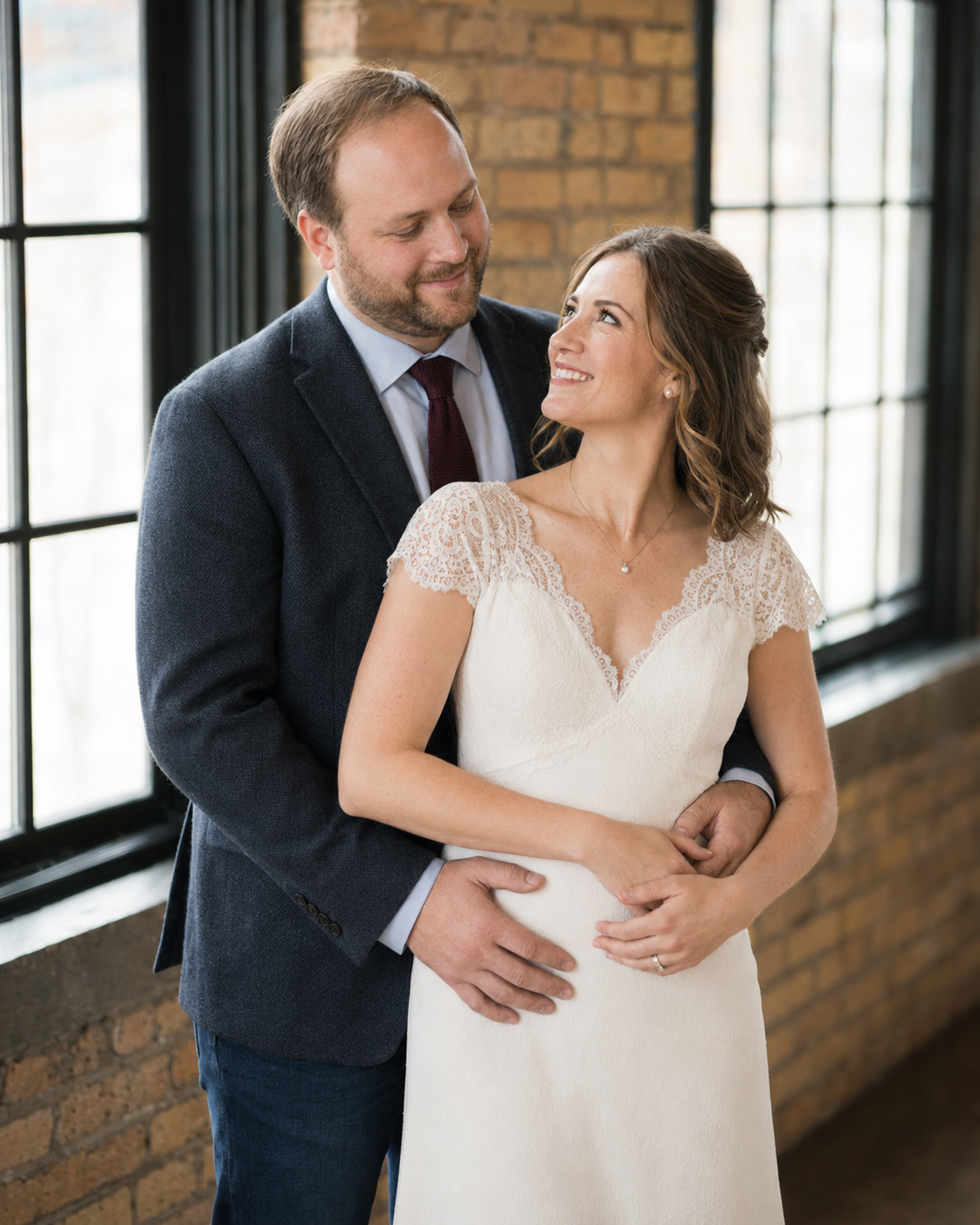 A smiling bride in a lace wedding gown embraces her groom in a navy suit near brick walls and windows.