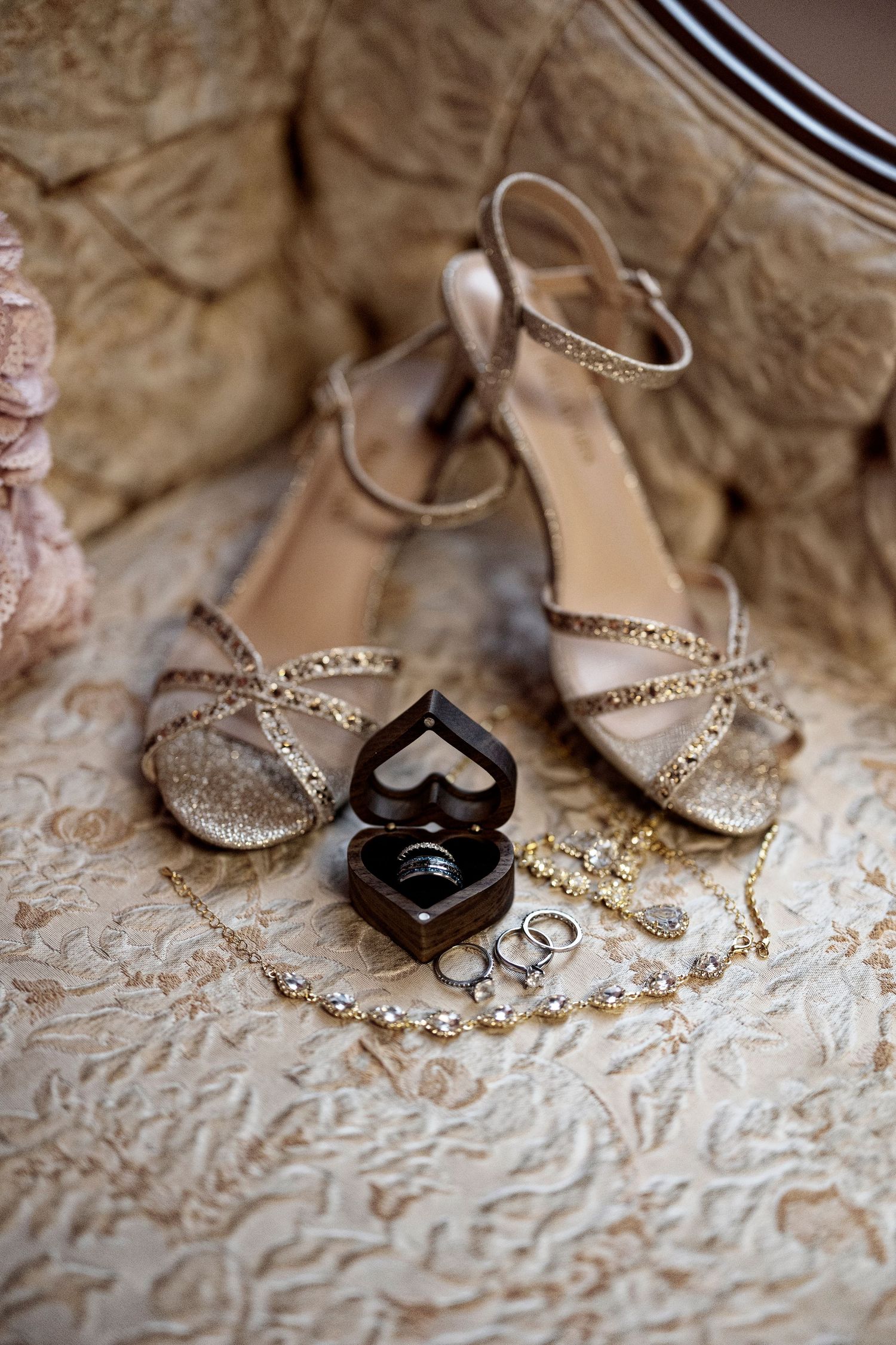 Gold strappy wedding heels beside a heart-shaped ring box with wedding bands and delicate jewelry on elegant fabric.