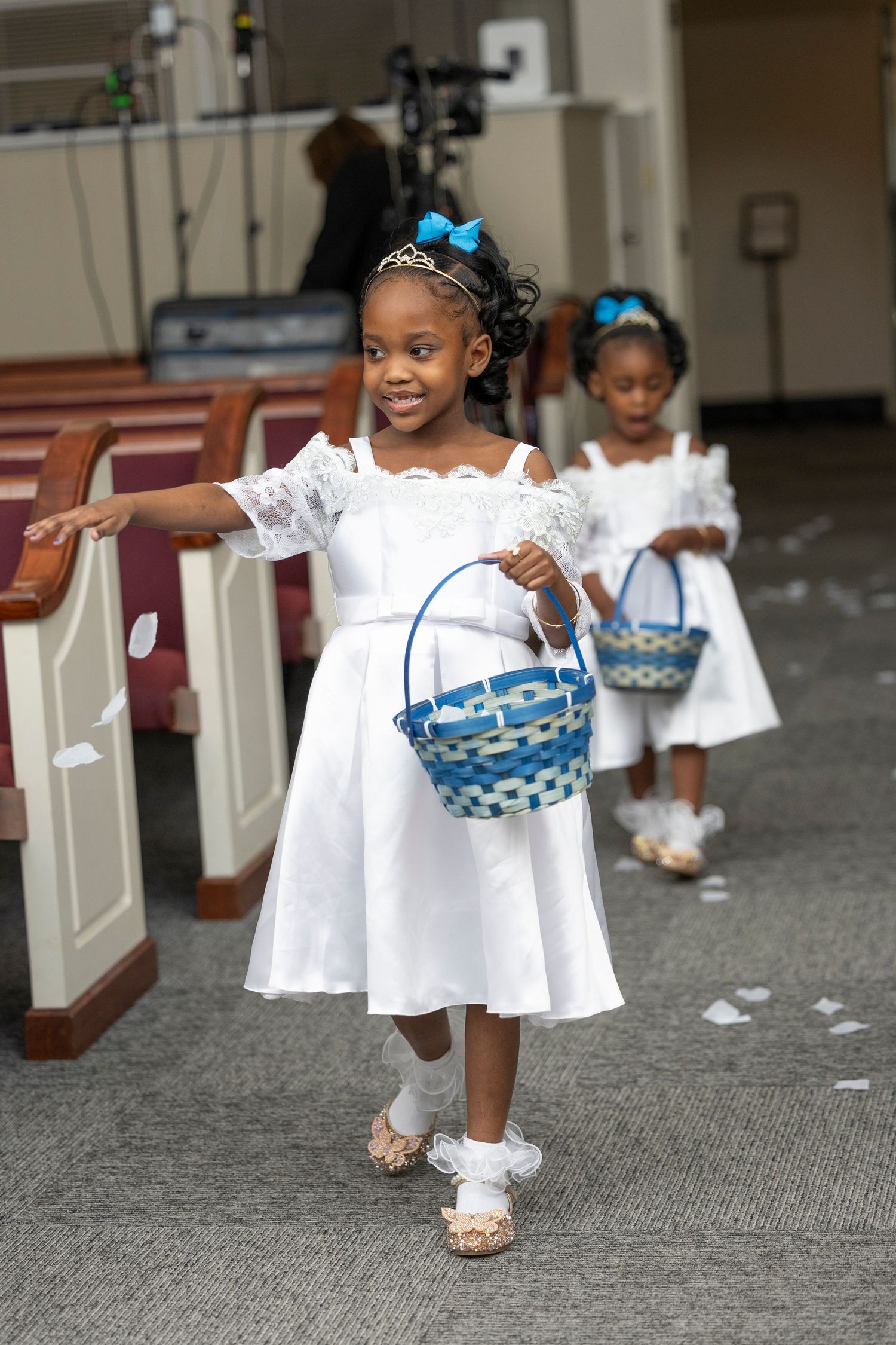 Two young flower girls in white dresses carry blue baskets down a church aisle at a wedding ceremony.