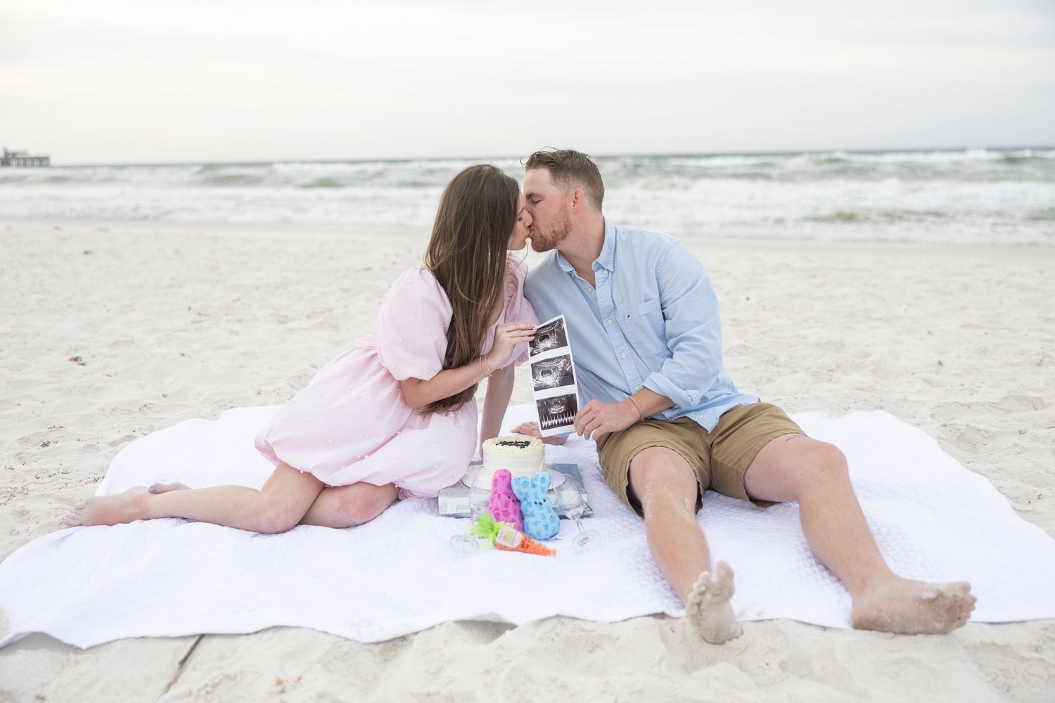Couple kissing on beach blanket during pregnancy announcement photoshoot with ultrasound photos and baby toys.