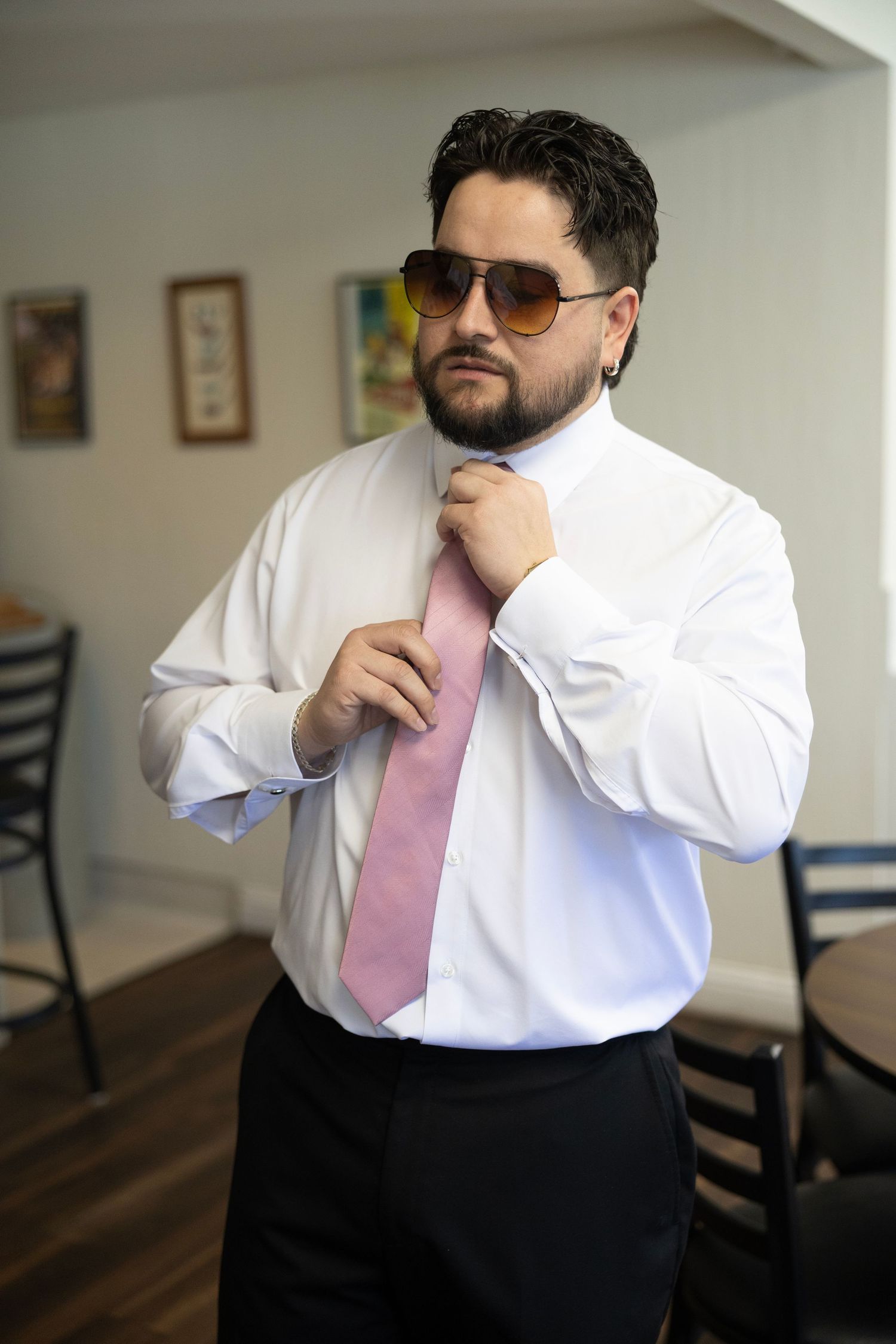 Man in white dress shirt and pink tie adjusting neckwear while wearing aviator sunglasses indoors.