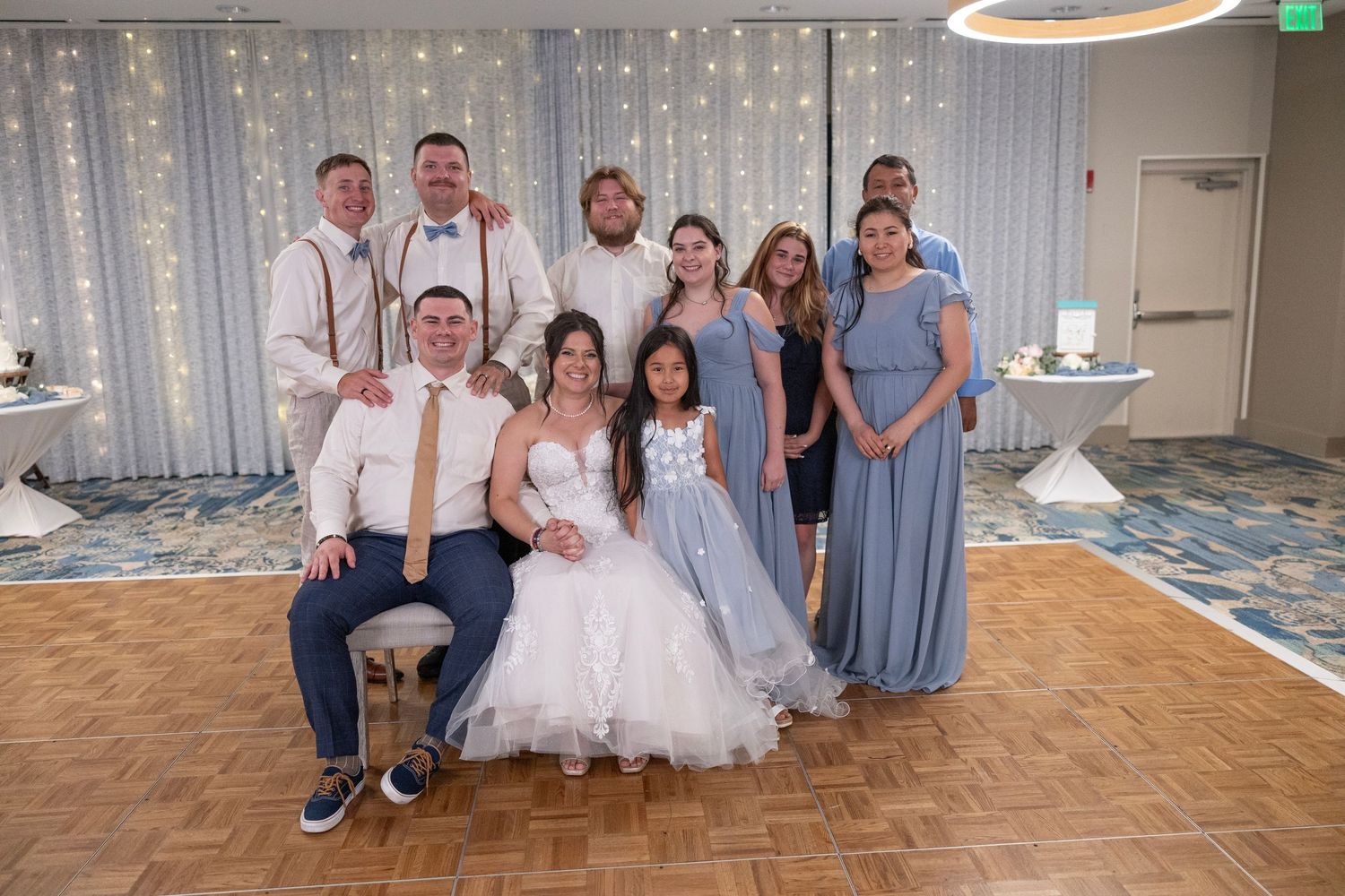 Wedding party poses together on dance floor with bride and groom seated in front, string lights backdrop.