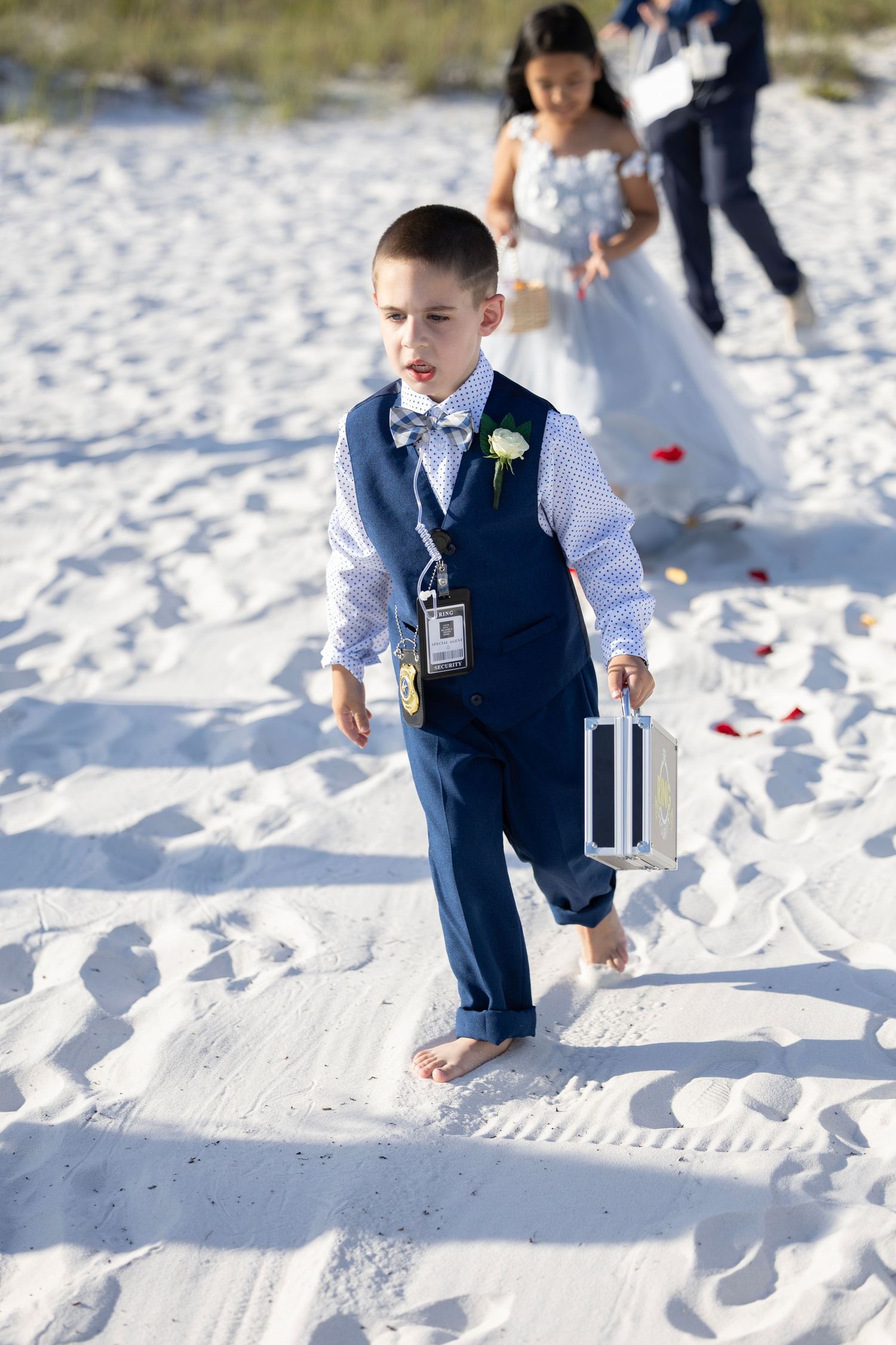 Young ring bearer in navy suit walks barefoot on white sand beach during outdoor wedding ceremony.