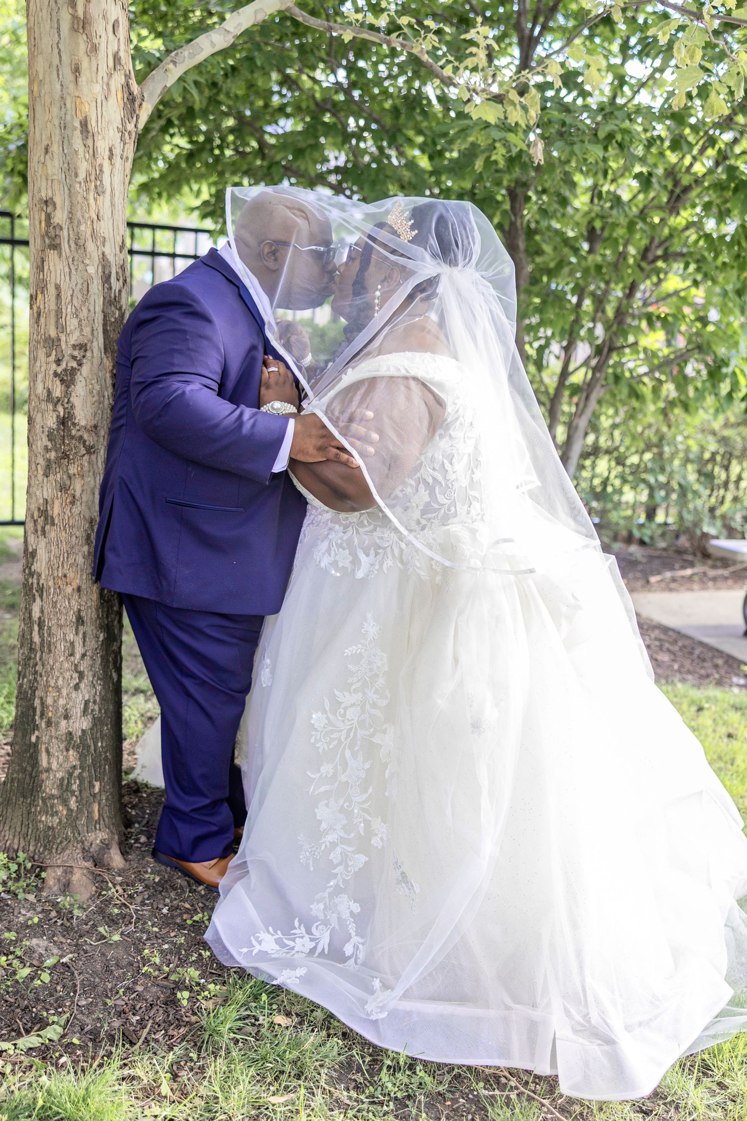 Newlywed couple sharing a romantic kiss under a veil outdoors, groom in blue suit, bride in white lace gown.