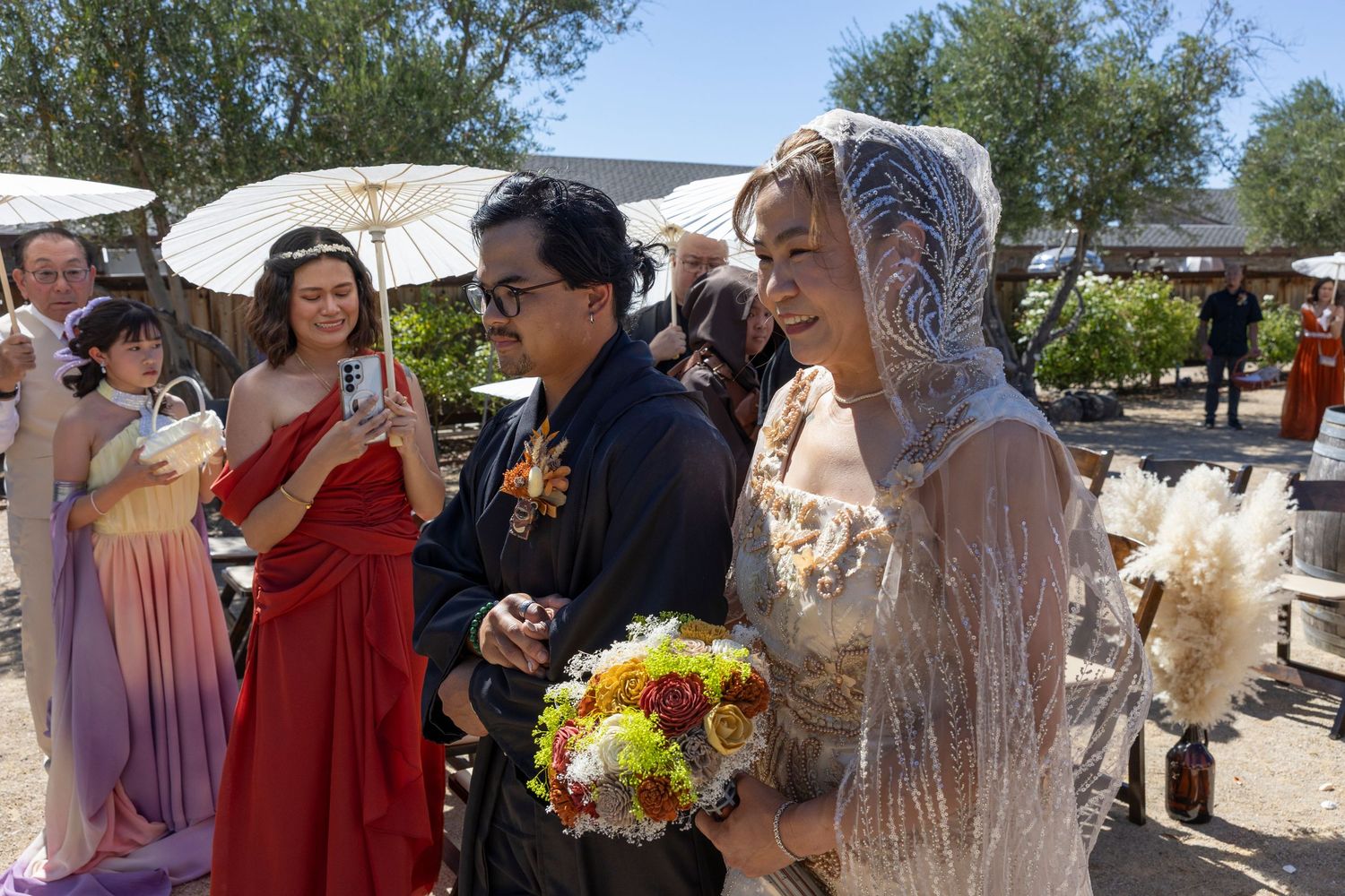 Bride in lace veil and groom in black suit walk together outdoors at a wedding ceremony with guests.
