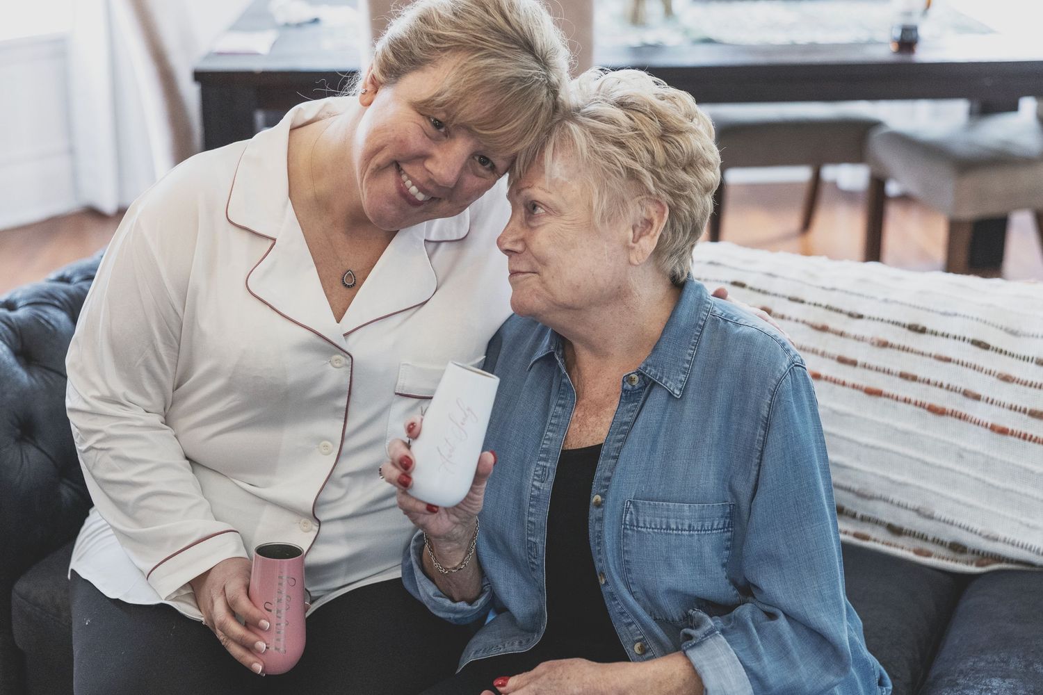 Two women sitting together on a couch, sharing a warm moment while holding mugs in a cozy living room.