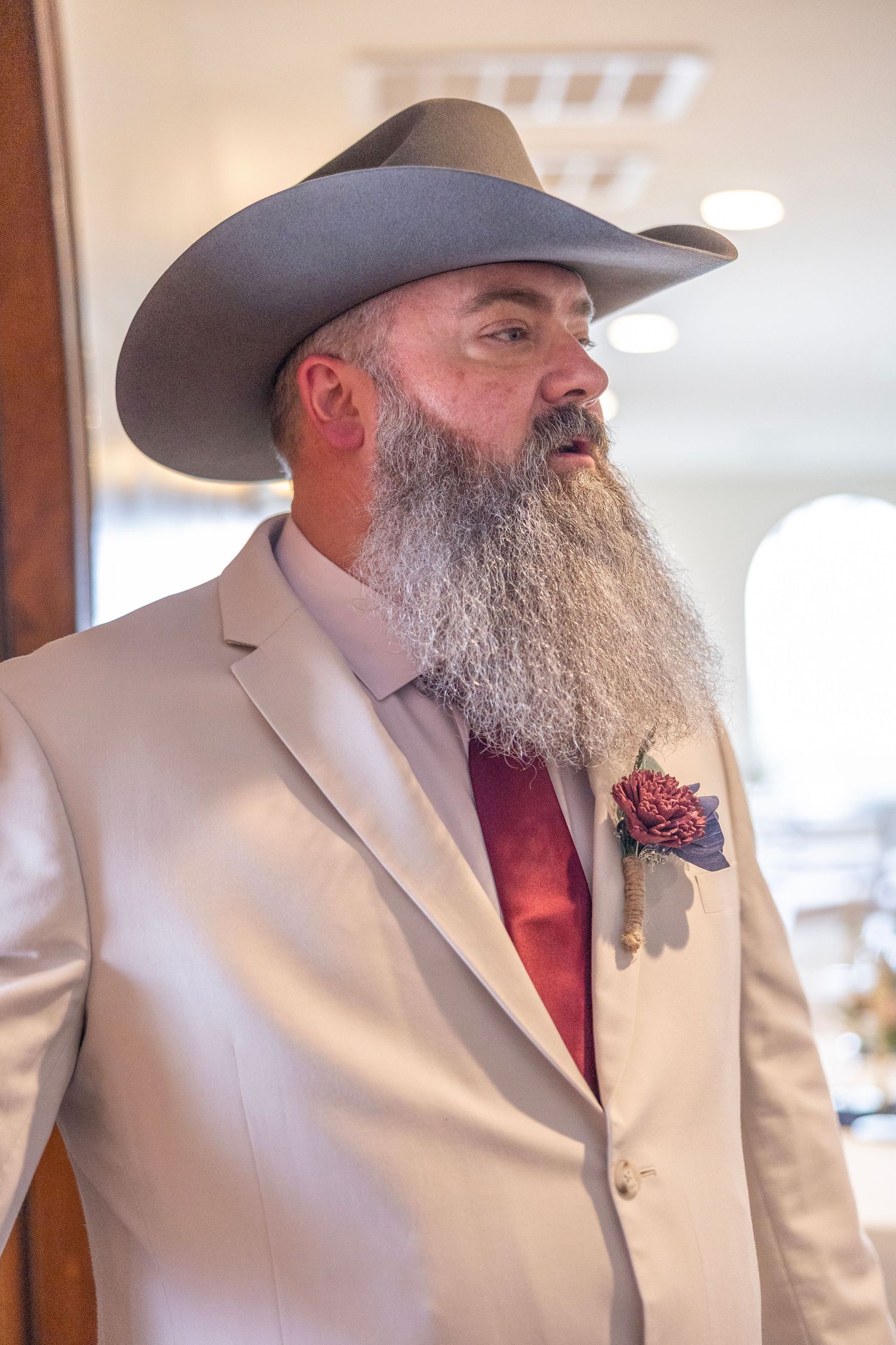 Bearded man in white suit, red tie, boutonniere, and gray cowboy hat at a formal event.