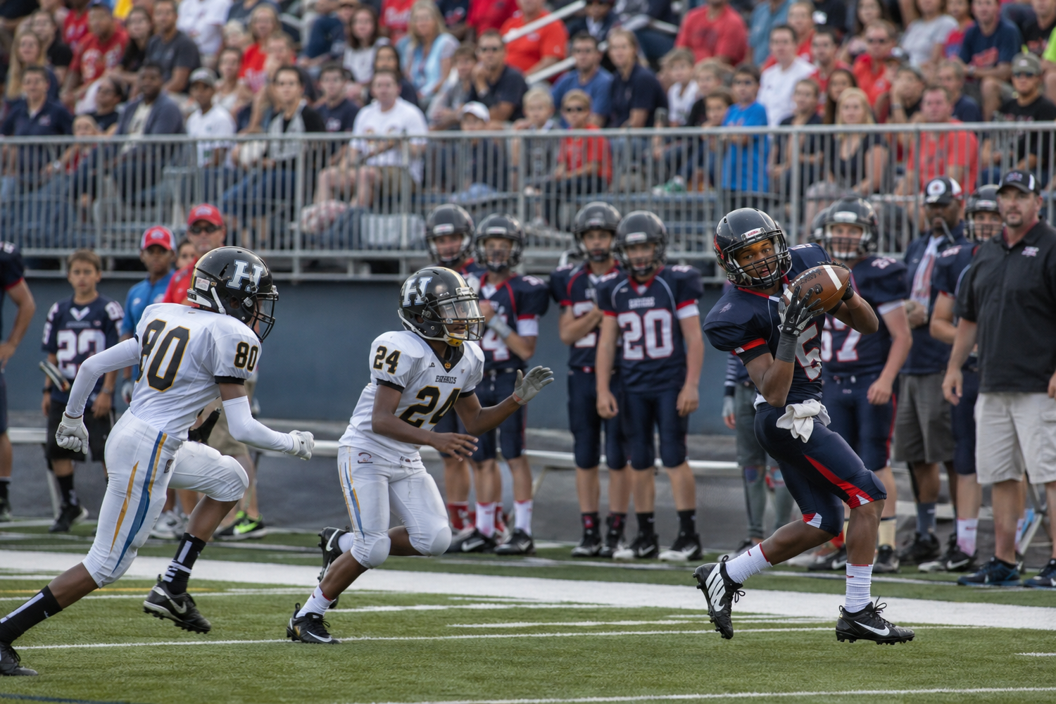 Football player in navy uniform evades two defenders in gold jerseys during a competitive game.