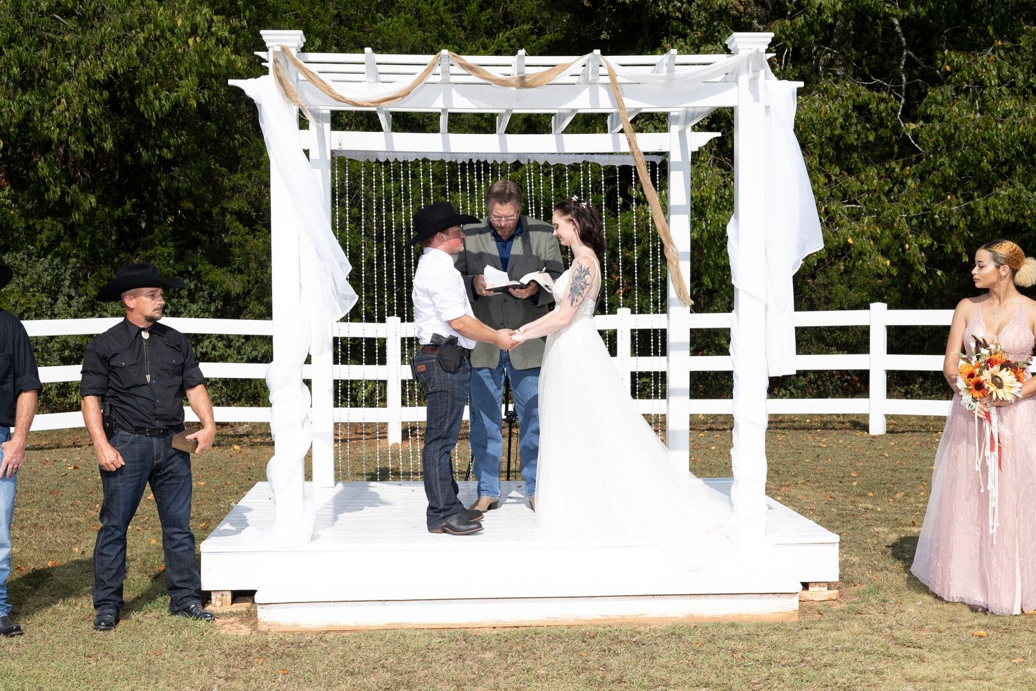 Wedding ceremony under white pergola with attendees gathered around couple taking vows.