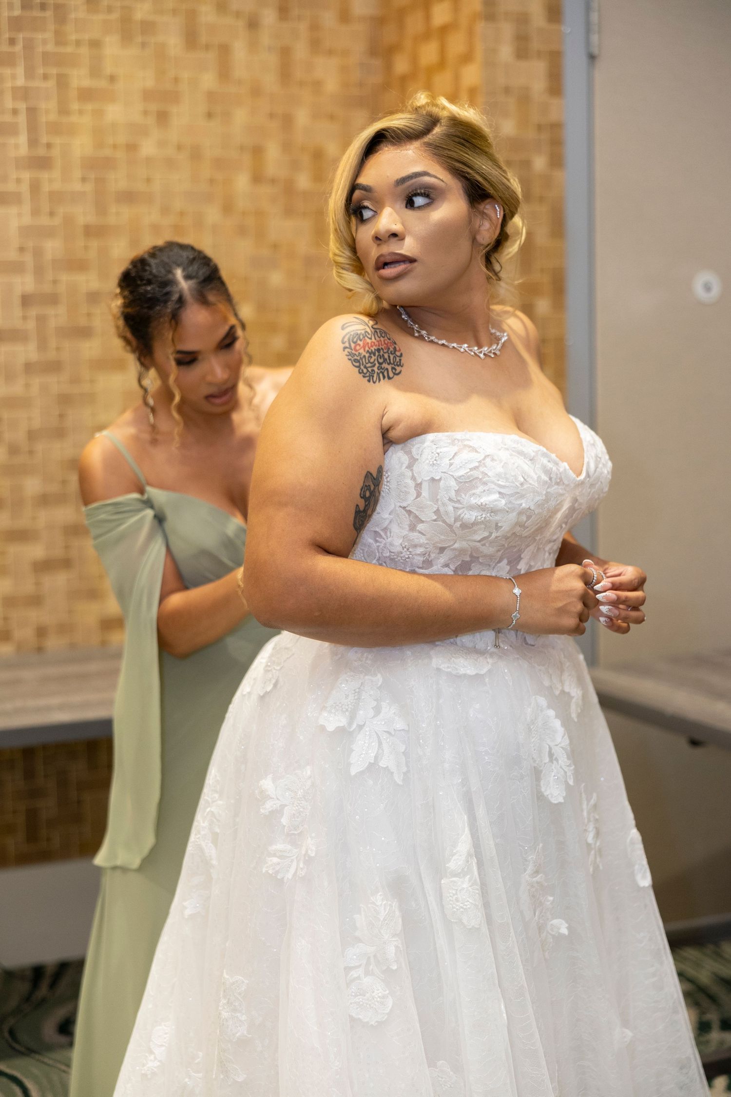 Bride in a flowing white lace wedding gown gets help with final adjustments while bridesmaid in sage green dress assists.