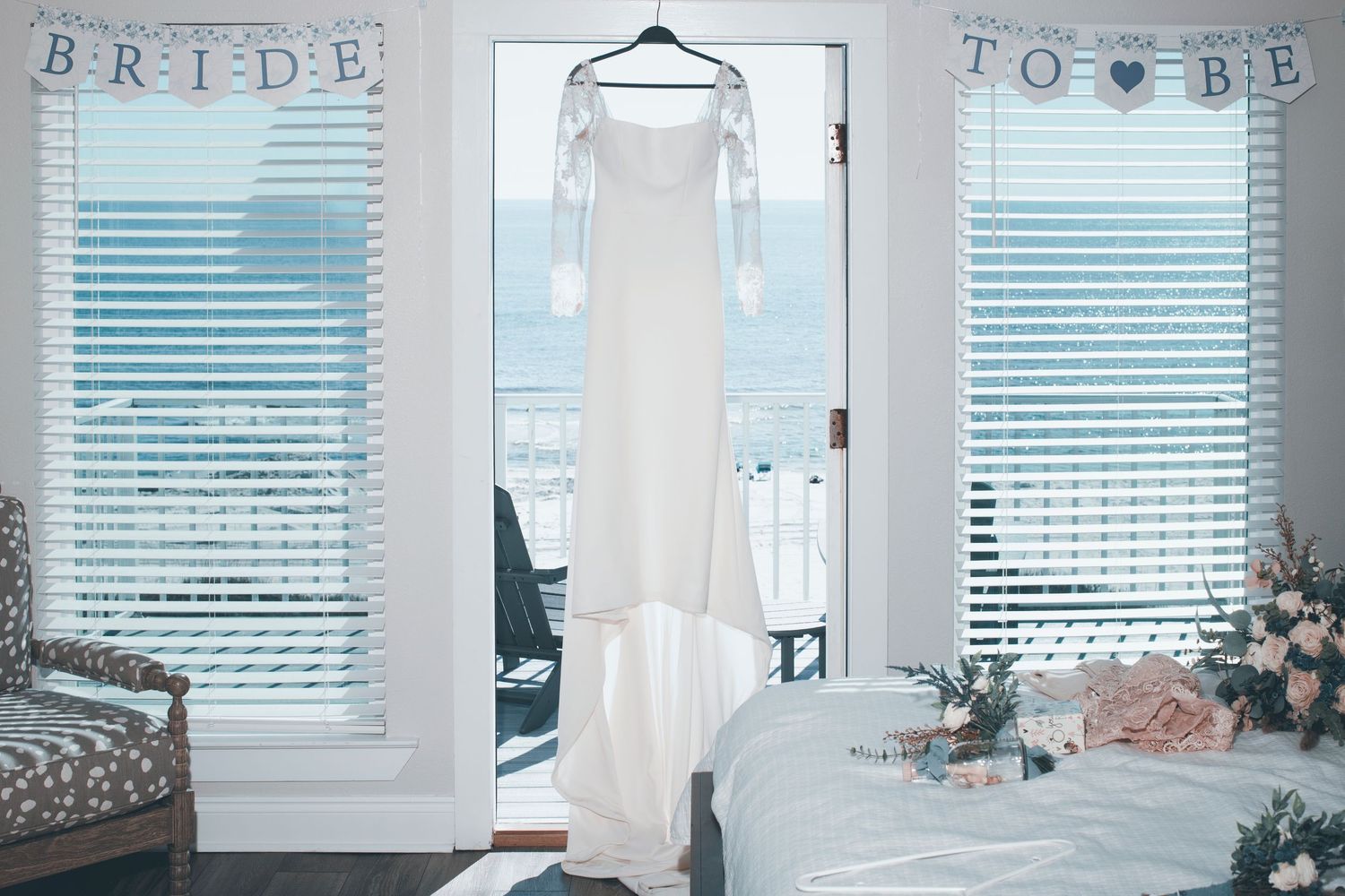 A sleek white wedding dress hangs in a bridal suite with blue tinted windows and 'BRIDE TO BE' banner decoration.