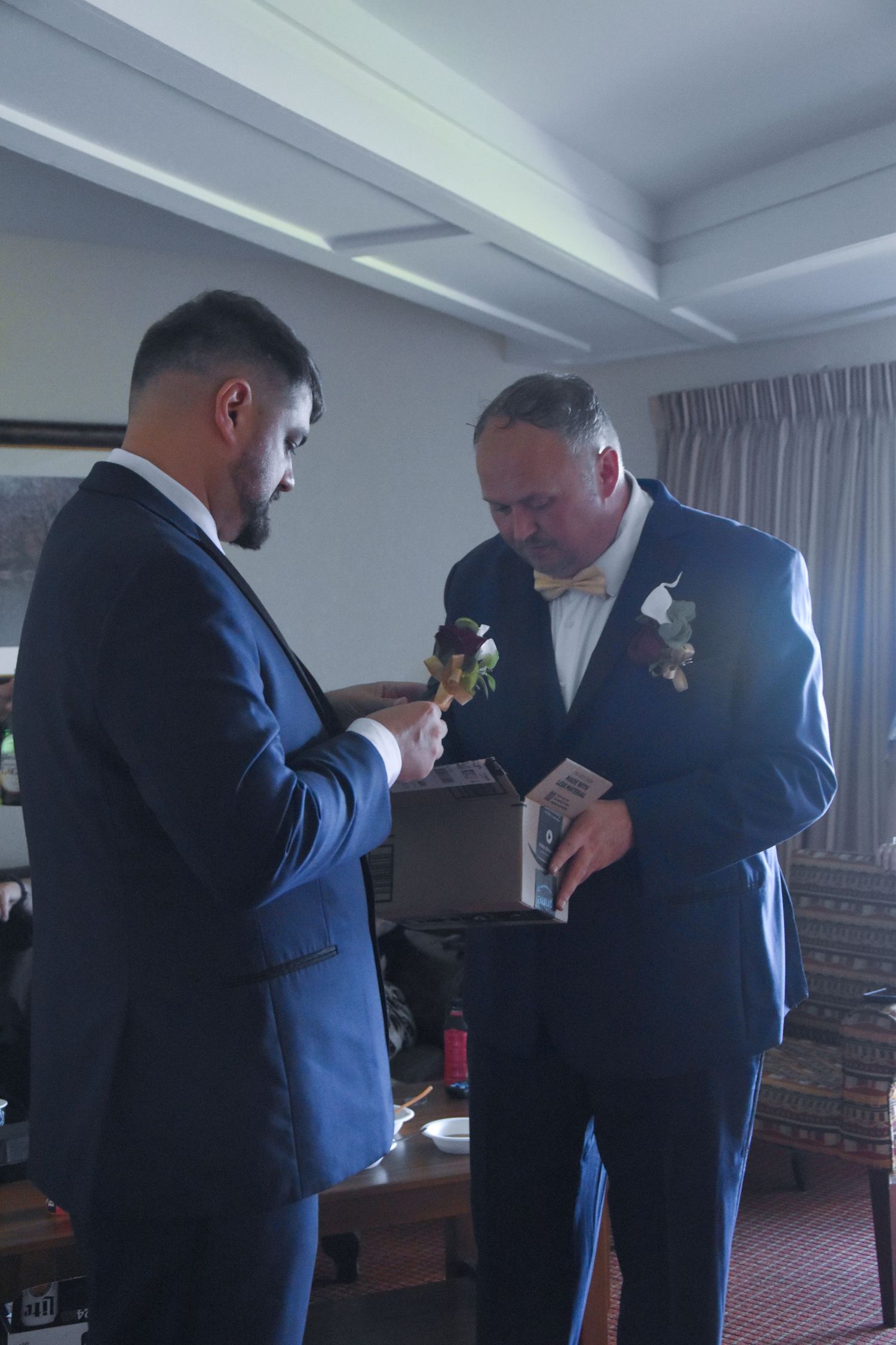 Two men in navy suits adjust their ties and boutonnieres while getting ready for a formal event.