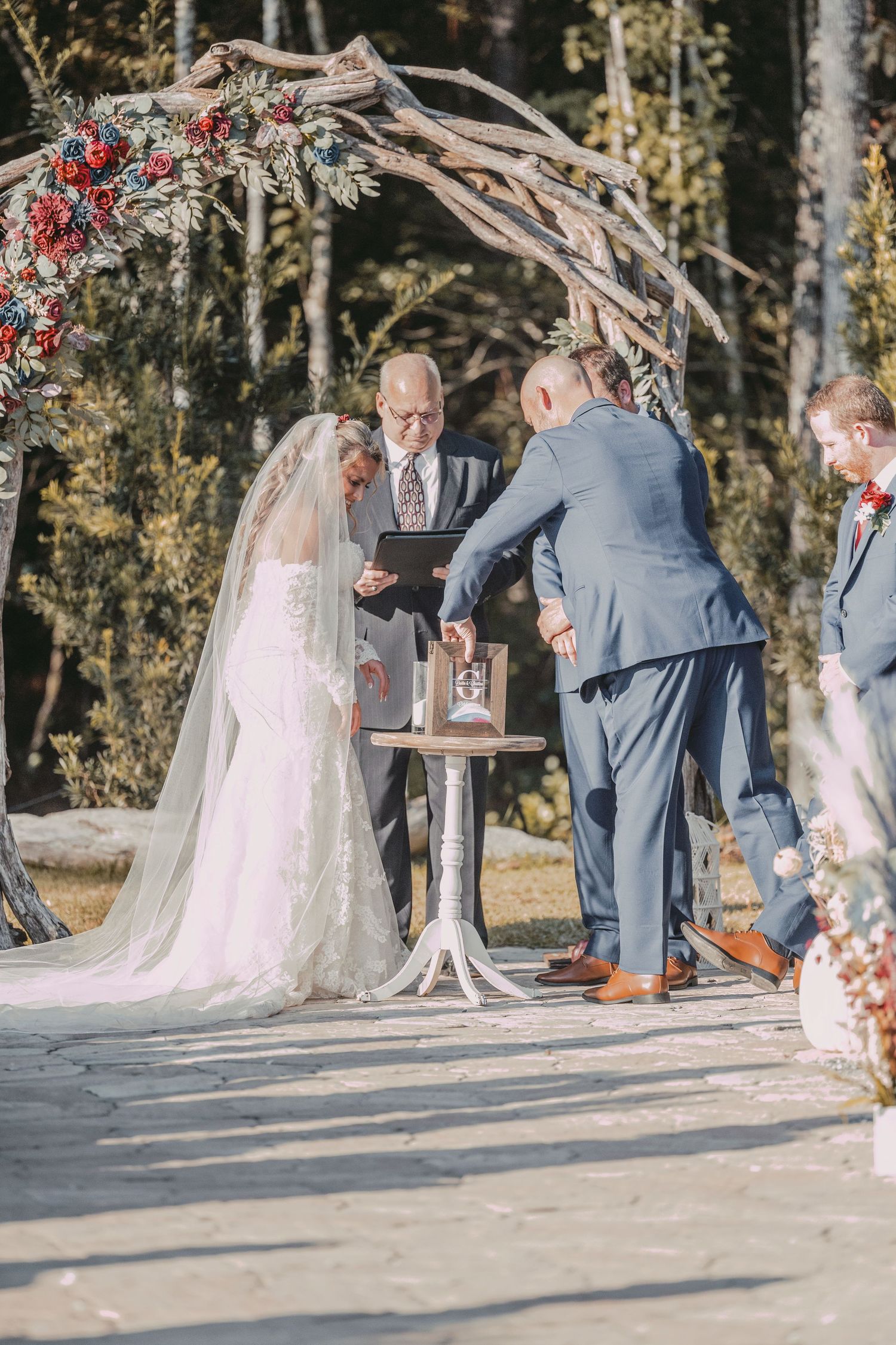 A wedding ceremony takes place under a rustic arch decorated with red flowers and greenery in an outdoor woodland setting.