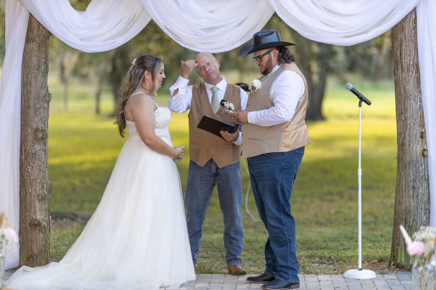 Outdoor wedding ceremony taking place under a white draped canopy in a sunny park setting.