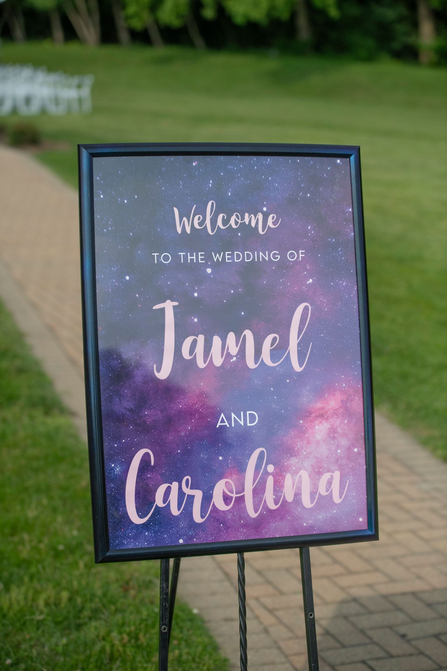 Wedding welcome sign with galaxy-themed purple and blue watercolor background displayed along a brick pathway.