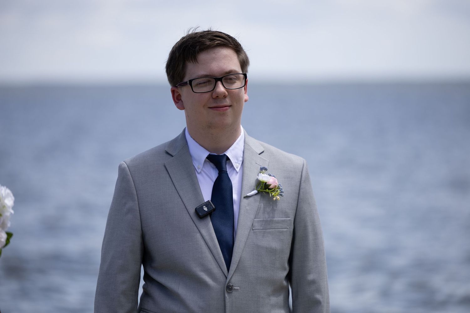 A groom in a light gray suit and navy tie stands outdoors with a boutonniere on a cloudy day.