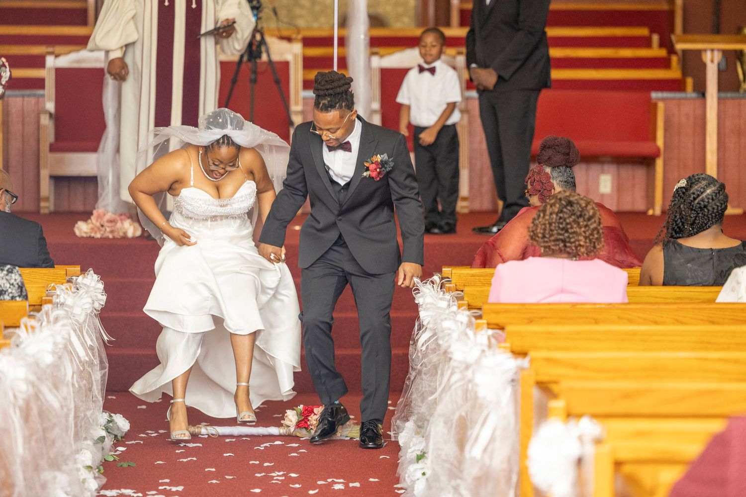 A couple walks down a church aisle decorated with white and red decor during a wedding ceremony.