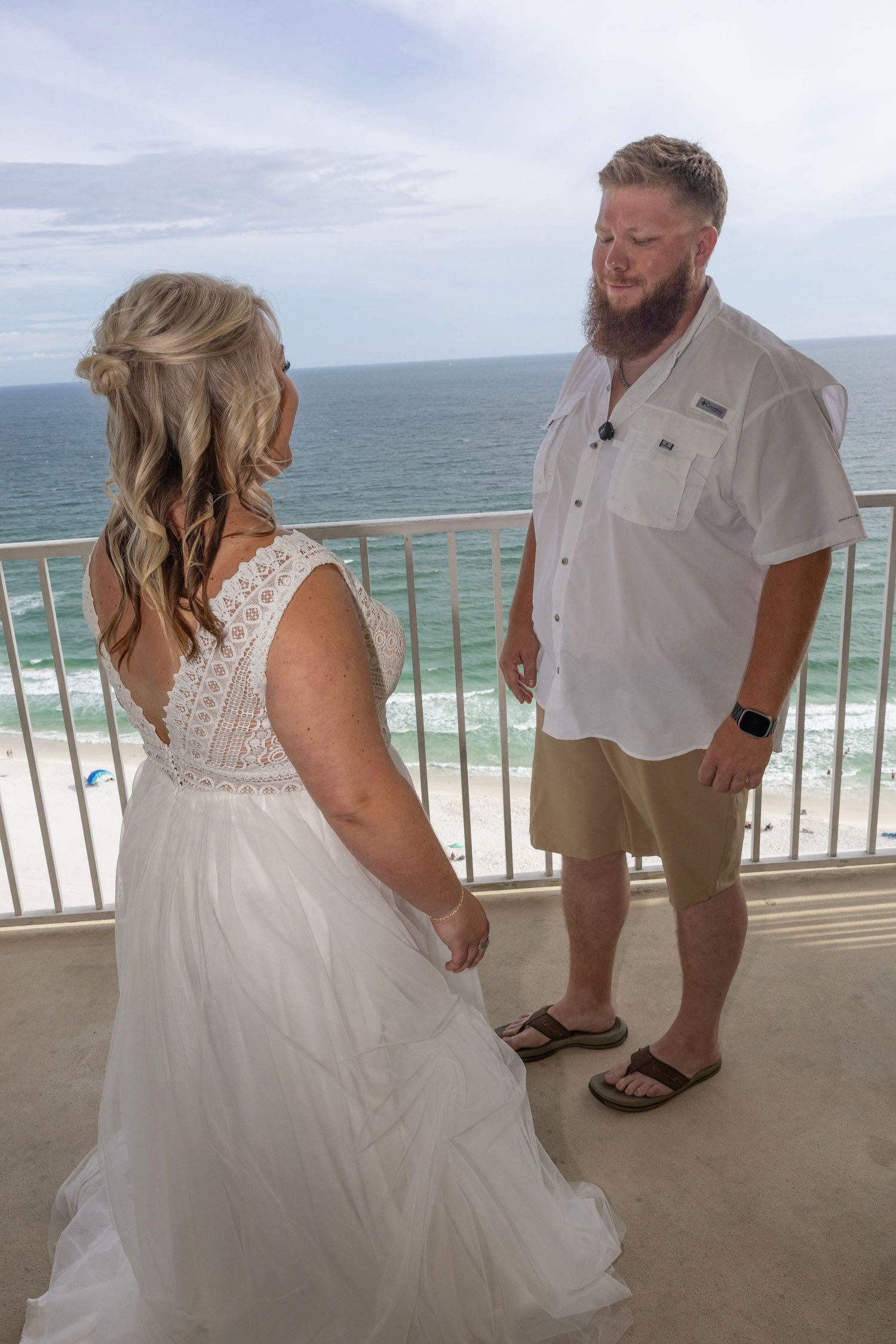 Bride in white lace gown shares first look moment with bearded groom on ocean-view balcony.