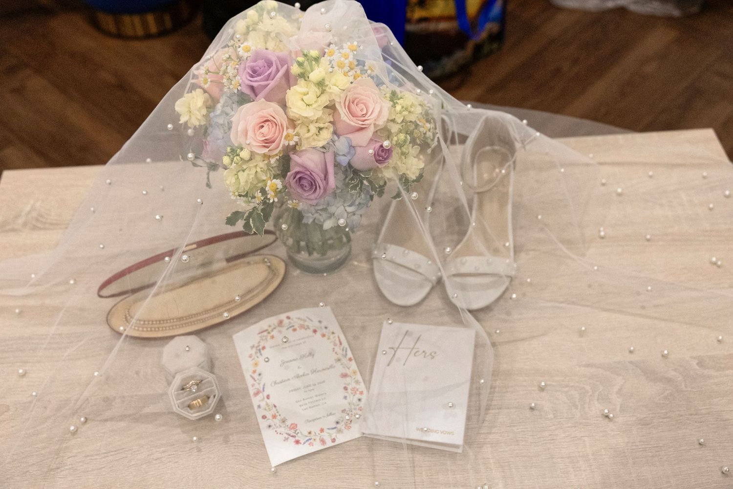 Elegant wedding flat lay with pastel floral bouquet, bridal heels, veil, rings, and invitation cards on a wooden table.