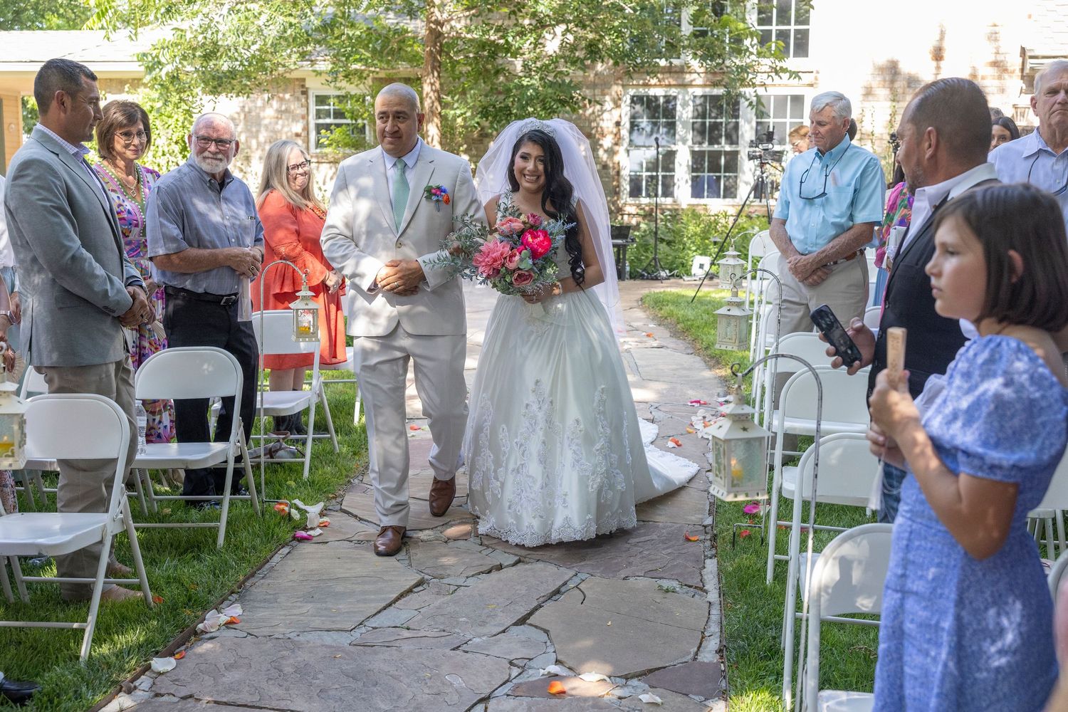 Bride in white gown walks down outdoor aisle with escort, flanked by seated guests at a sunny garden wedding ceremony.