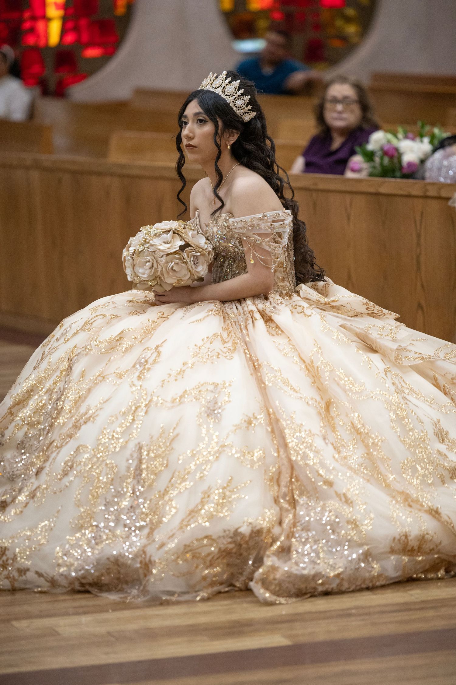 Young woman in gold quinceañera ball gown with tiara holding floral bouquet during church ceremony.