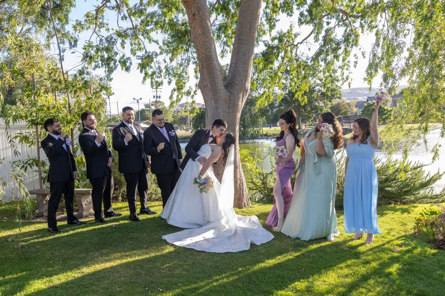 Wedding party celebrating outdoors under a large tree by a lake, with groomsmen and bridesmaids cheering the kissing couple.