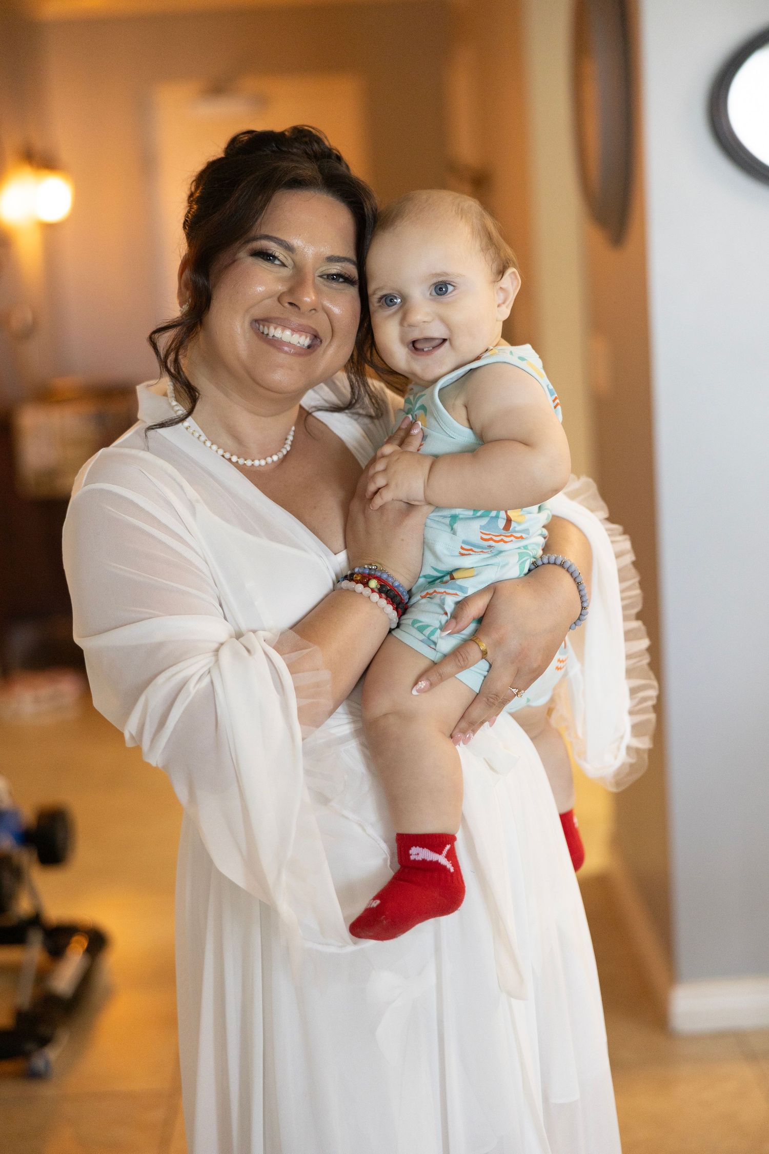A smiling woman in a white dress holds a happy baby in a floral outfit with red socks indoors.