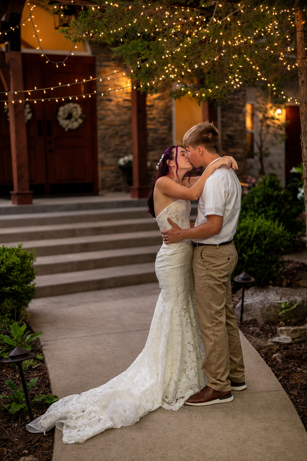 Bride and groom share a romantic kiss outdoors under string lights at their evening wedding venue.