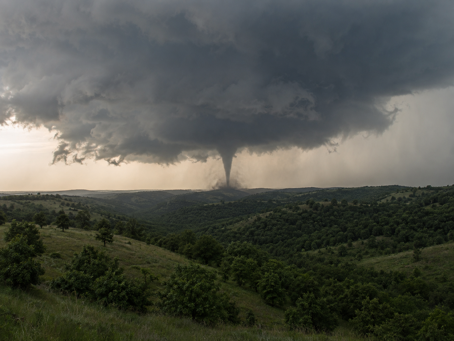 A massive tornado touches down over rolling green hills under dark storm clouds in a rural landscape.