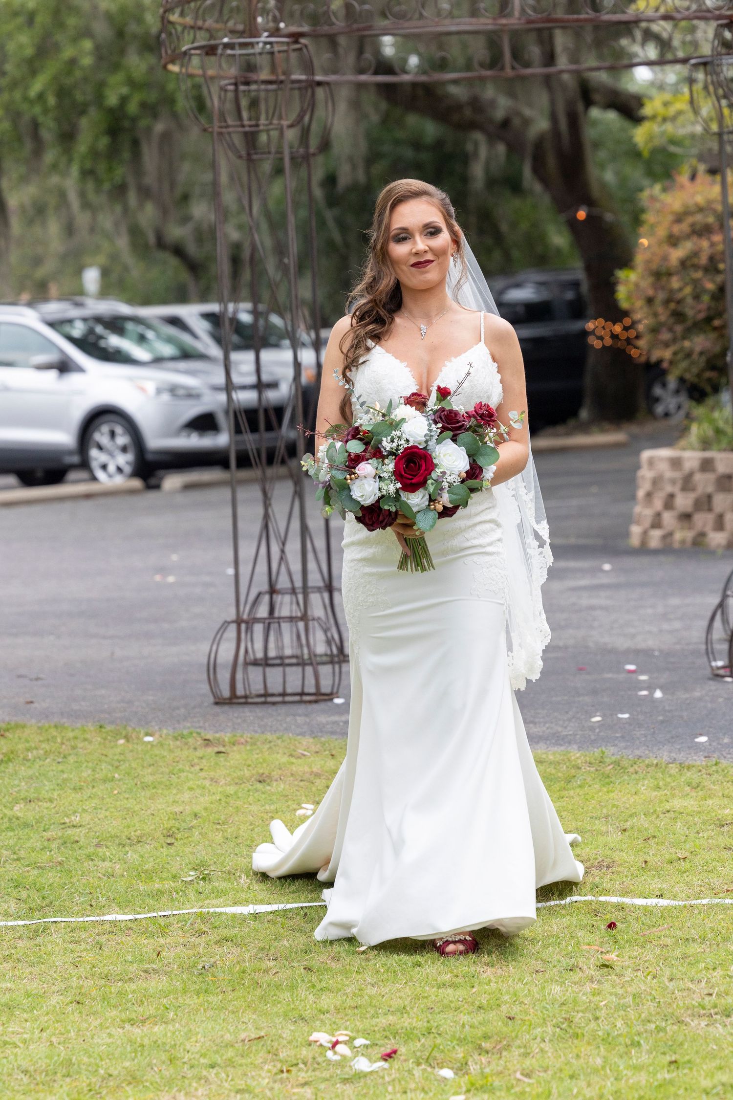A bride in a fitted white gown holds a burgundy and white bouquet while standing on grass near a metal arch.