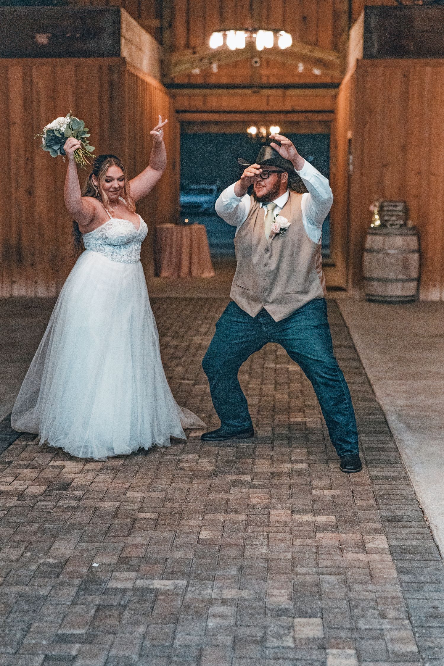 A fun wedding couple performs a choreographed dance sequence on a rustic brick floor in a barn venue.