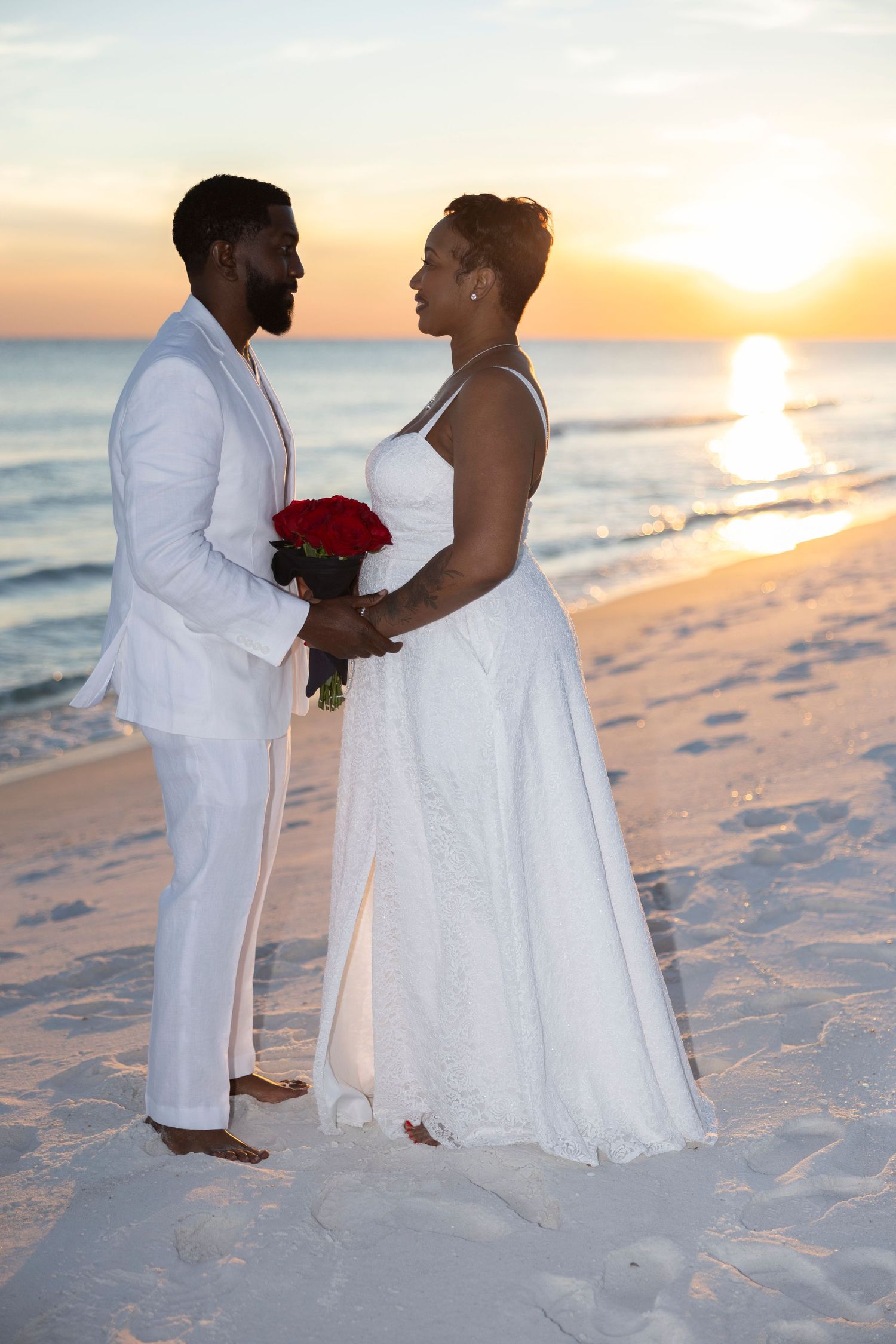 A romantic beach wedding photoshoot at sunset with a couple in white attire standing on pristine sand by the ocean.