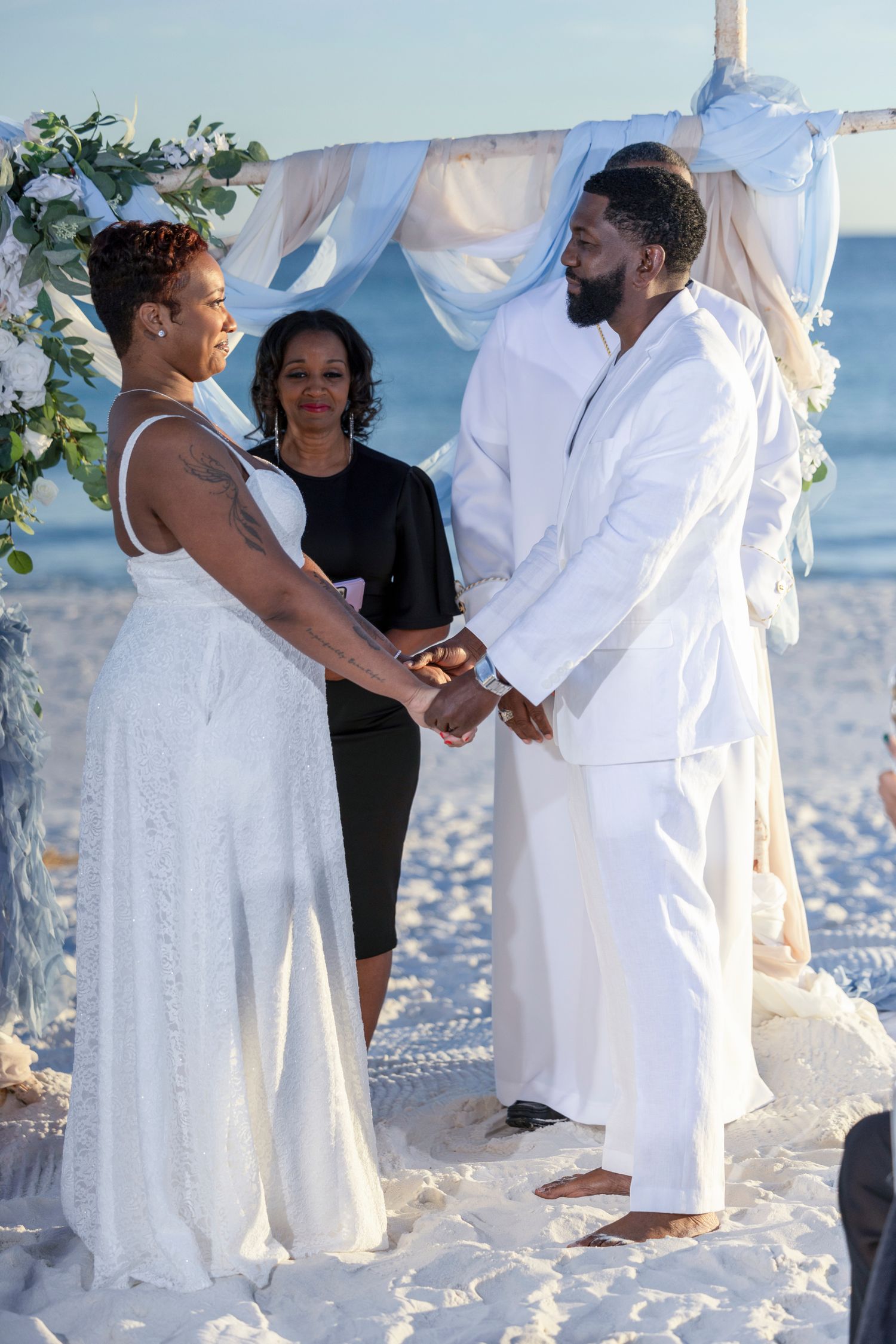 A romantic beach wedding ceremony takes place with the couple exchanging vows under an arch on white sandy shores.