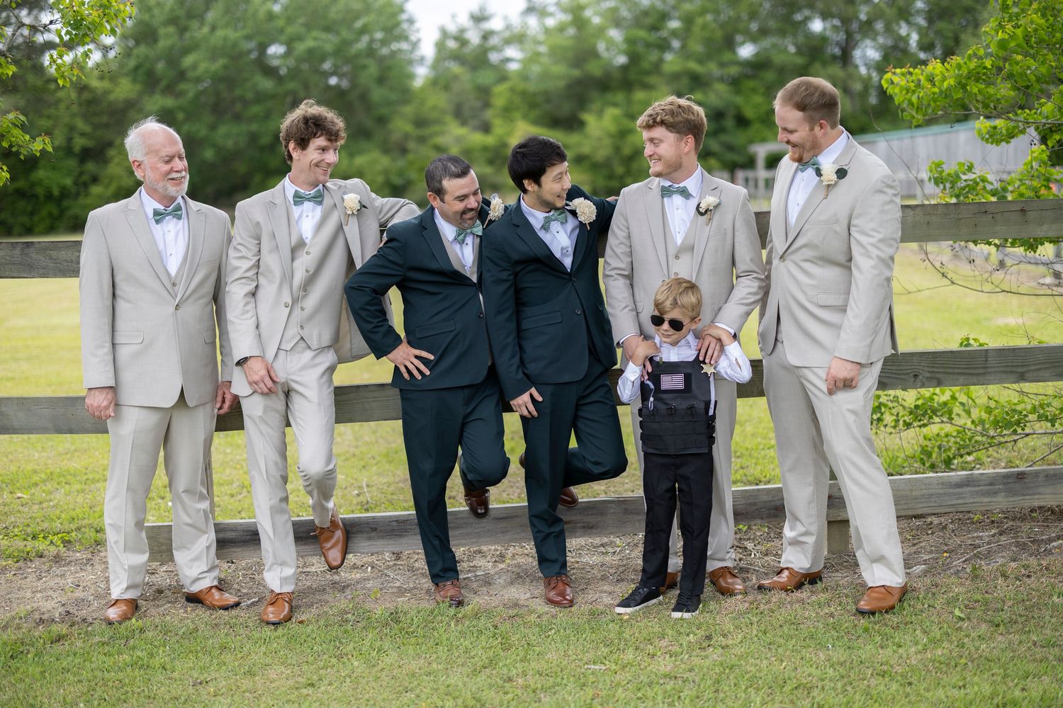 A group of groomsmen in light gray suits and dark suits pose casually on a grassy hillside for wedding photos.