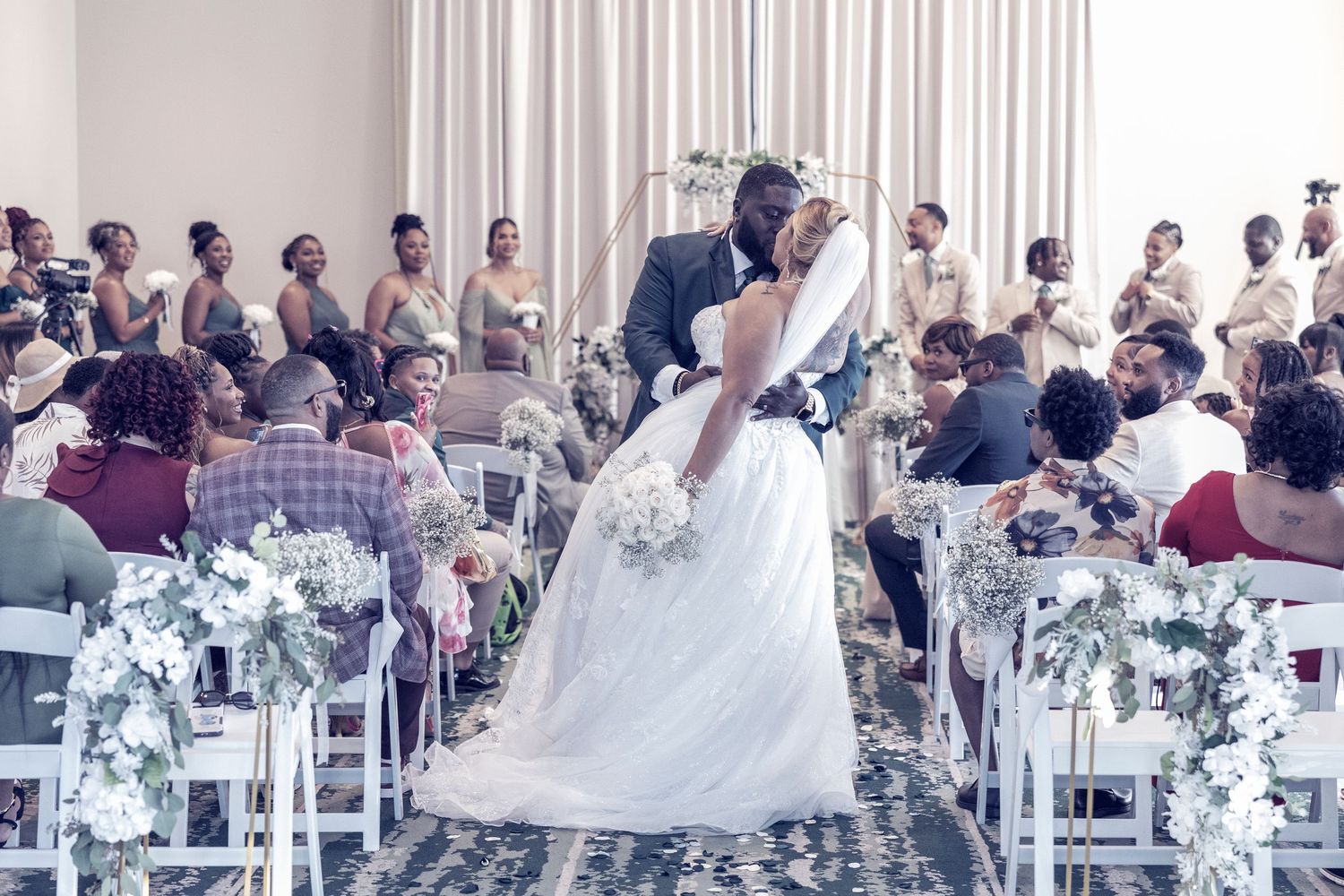 Wedding guests seated in white chairs with floral arrangements at an elegant indoor ceremony venue.