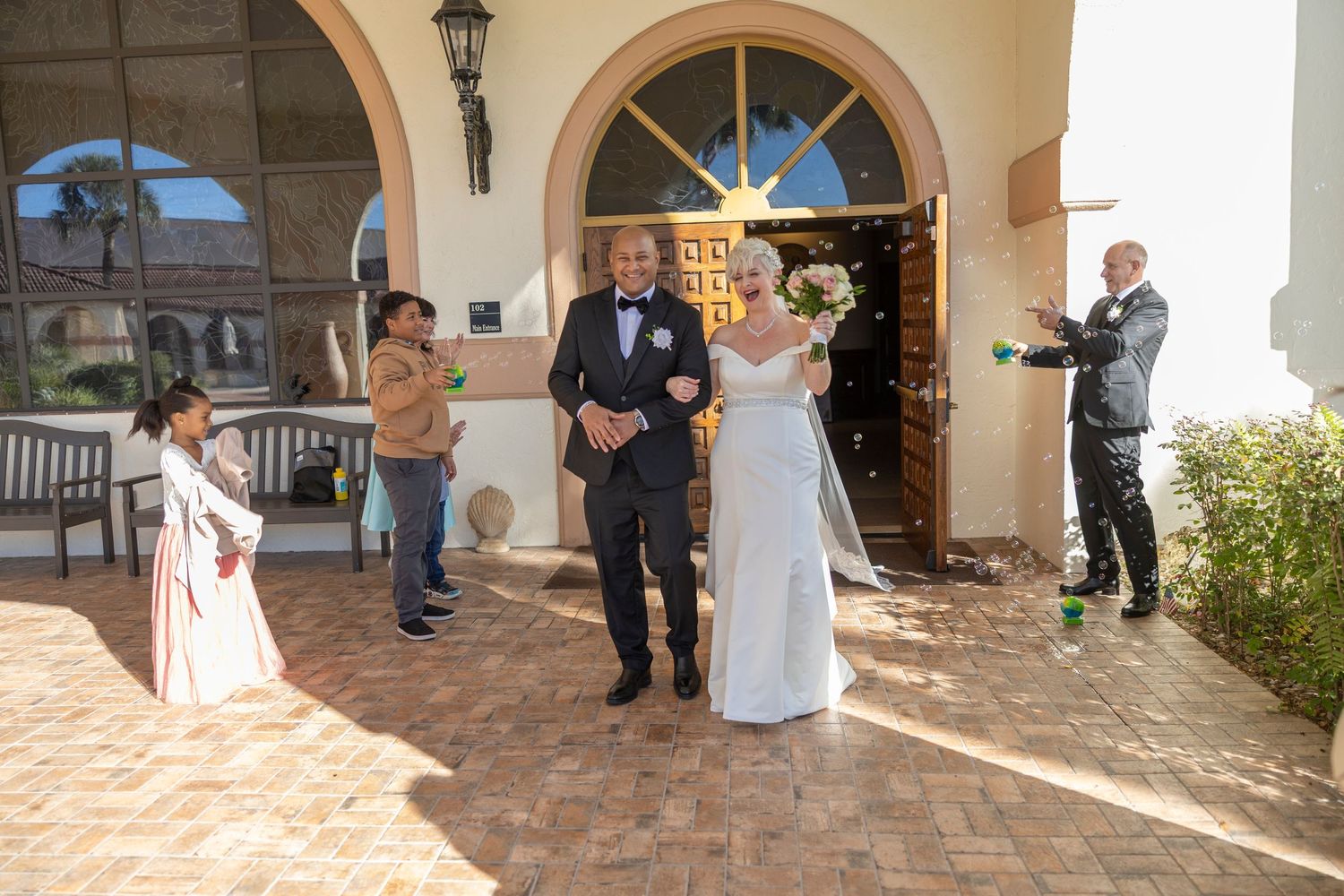 A wedding party gathers in an elegant Mediterranean-style courtyard with arched walkways and stone columns.