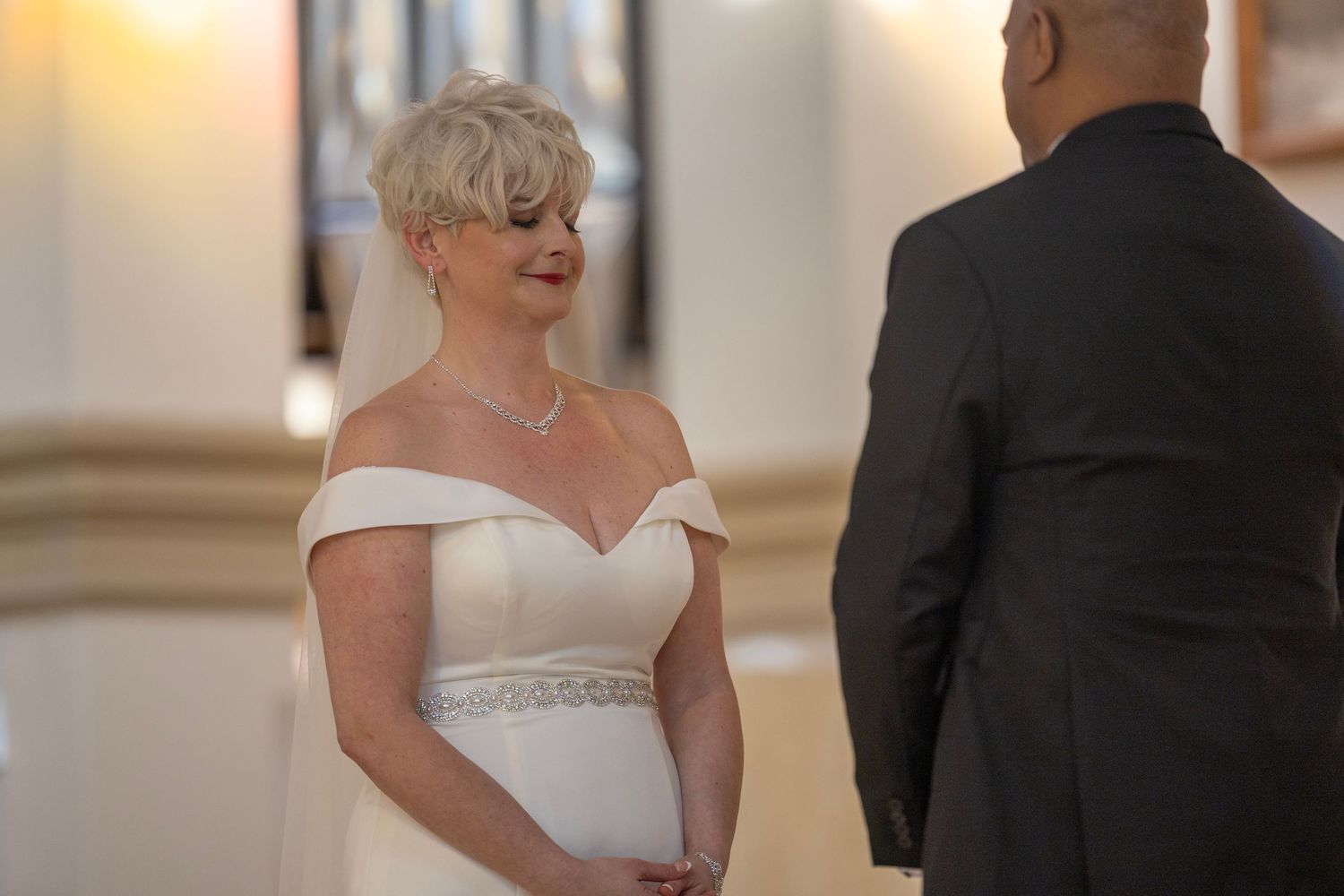 A bride in an elegant off-shoulder white gown with a beaded waistline smiles during a wedding ceremony.