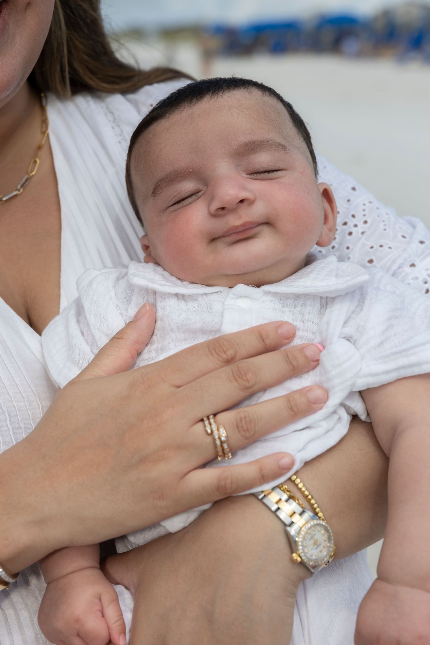 A sleeping baby wearing white clothing is gently touched by a hand wearing gold jewelry.